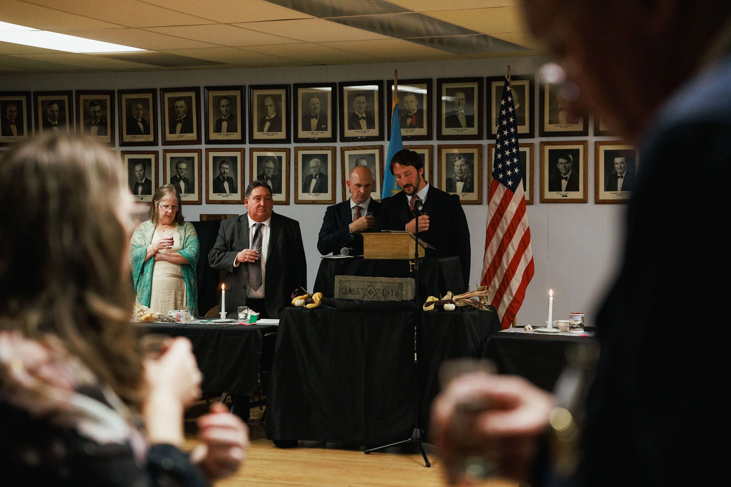 People gathered at a formal event, one man at a podium speaking into a microphone with an American flag behind him, framed photos of individuals on the wall in the background, and others standing and holding drinks.