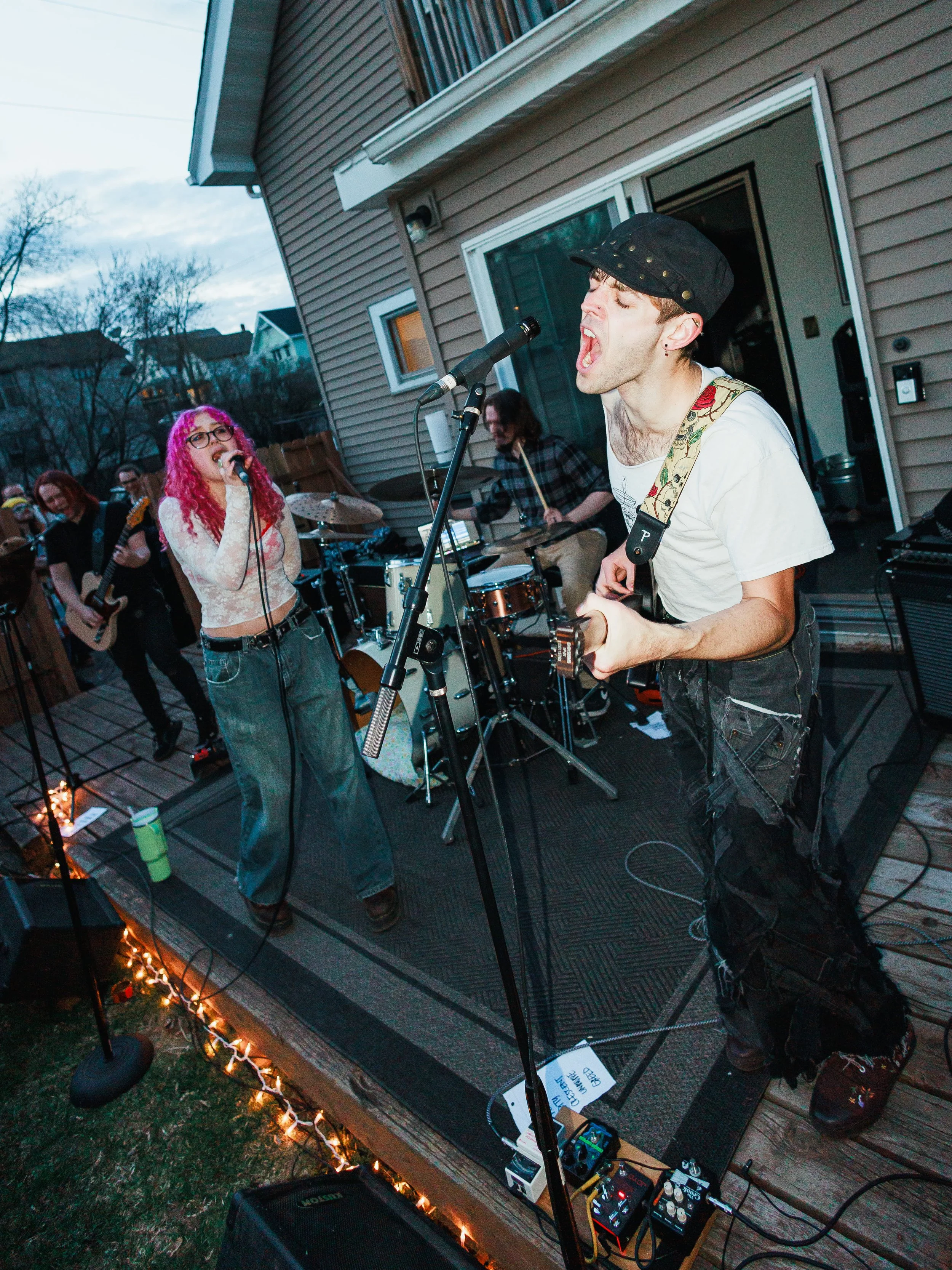 A group of musicians performing outdoors on a deck, including a woman with pink curly hair singing into a microphone, a man with a cap playing guitar and singing, and other band members playing drums and bass guitar, with string lights along the deck
