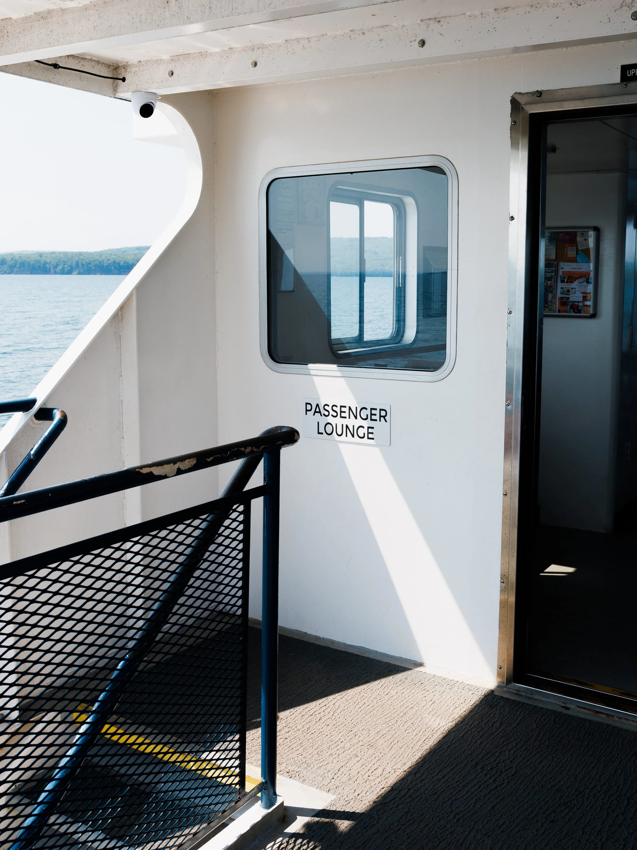 View of a Madeline Island ferry deck with a passageway leading to a passenger lounge, overlooking a body of water and distant land, captured during daylight.