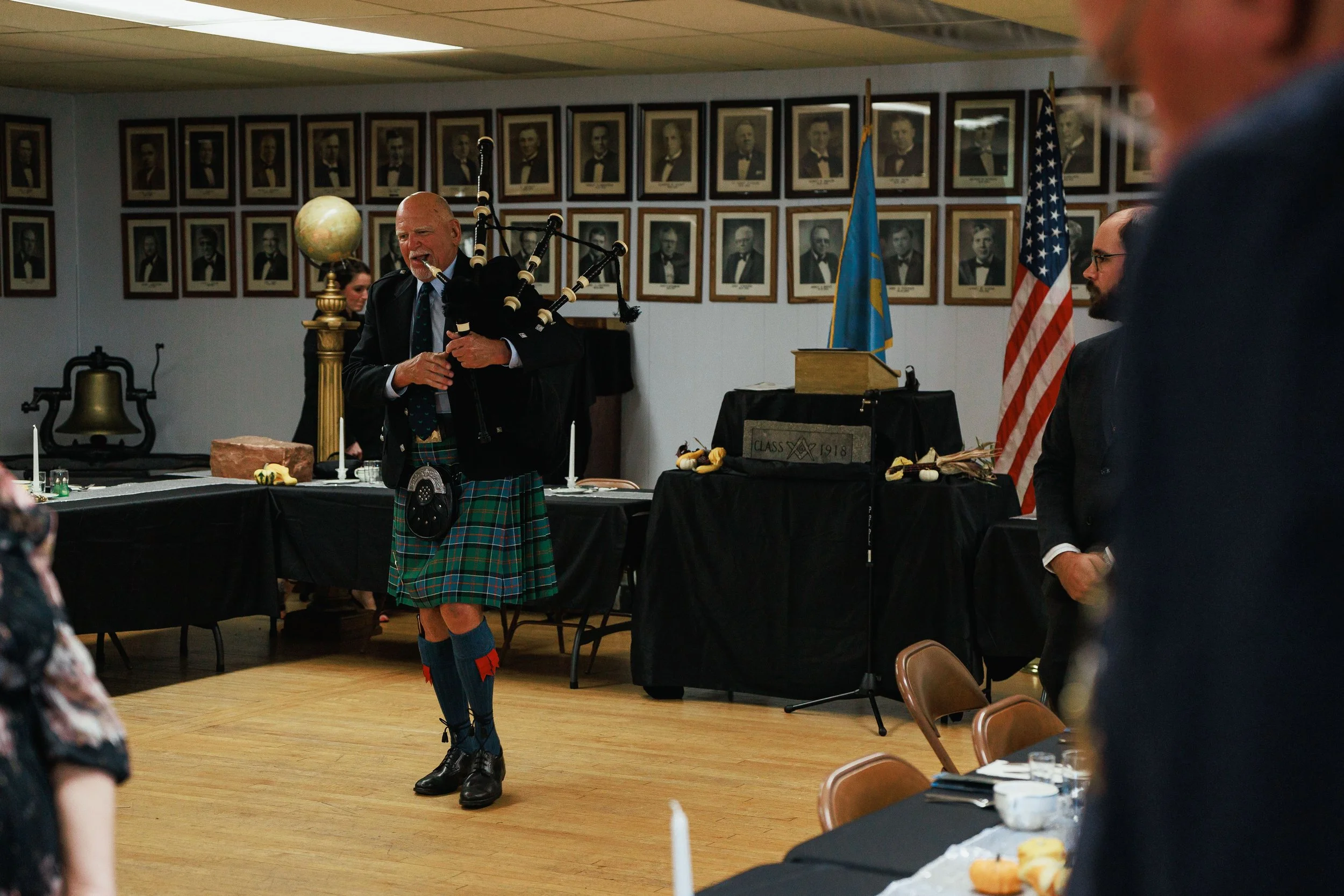 A man in a kilt playing bagpipes in a room decorated with framed photographs and flags.