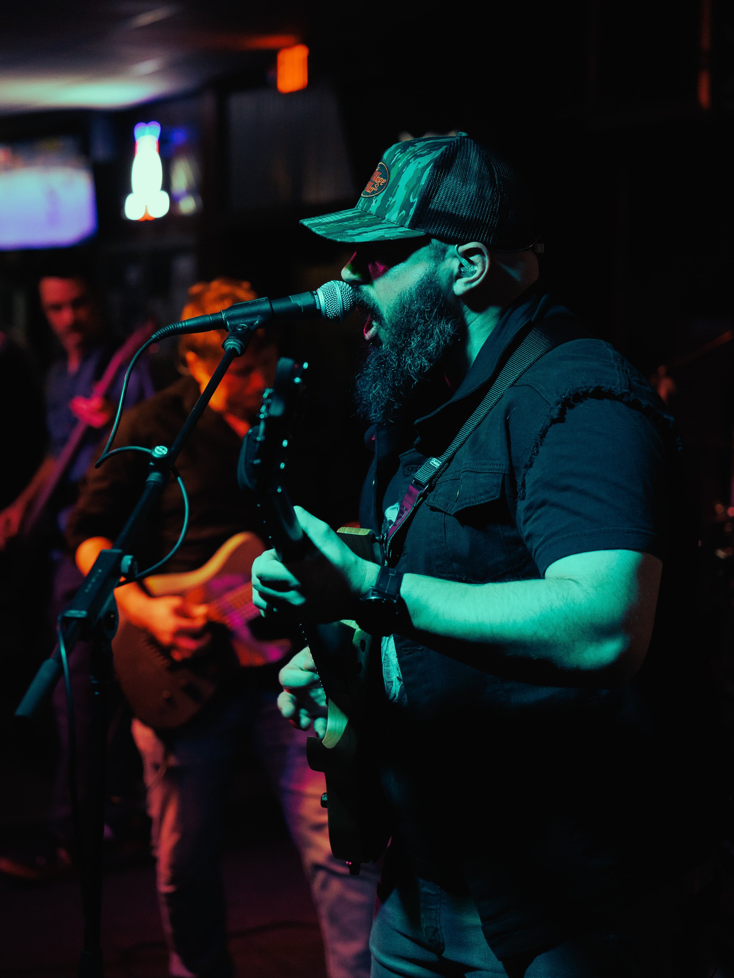 A musician with a beard singing into a microphone while playing an electric guitar on stage with other band members in the background.