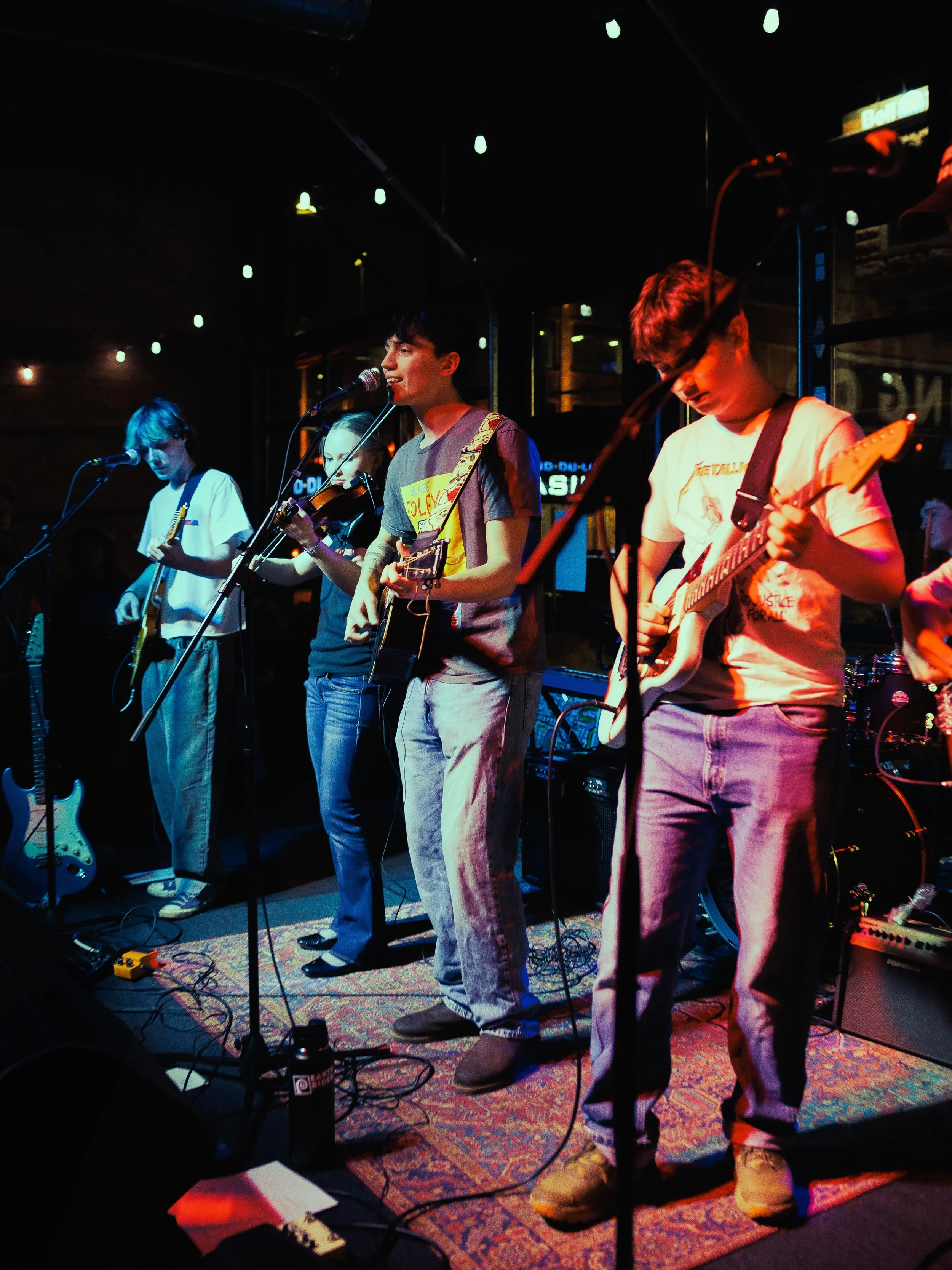 A band of four young musicians performing on stage at a small venue, with microphones and guitars, under dim, colorful lighting.
