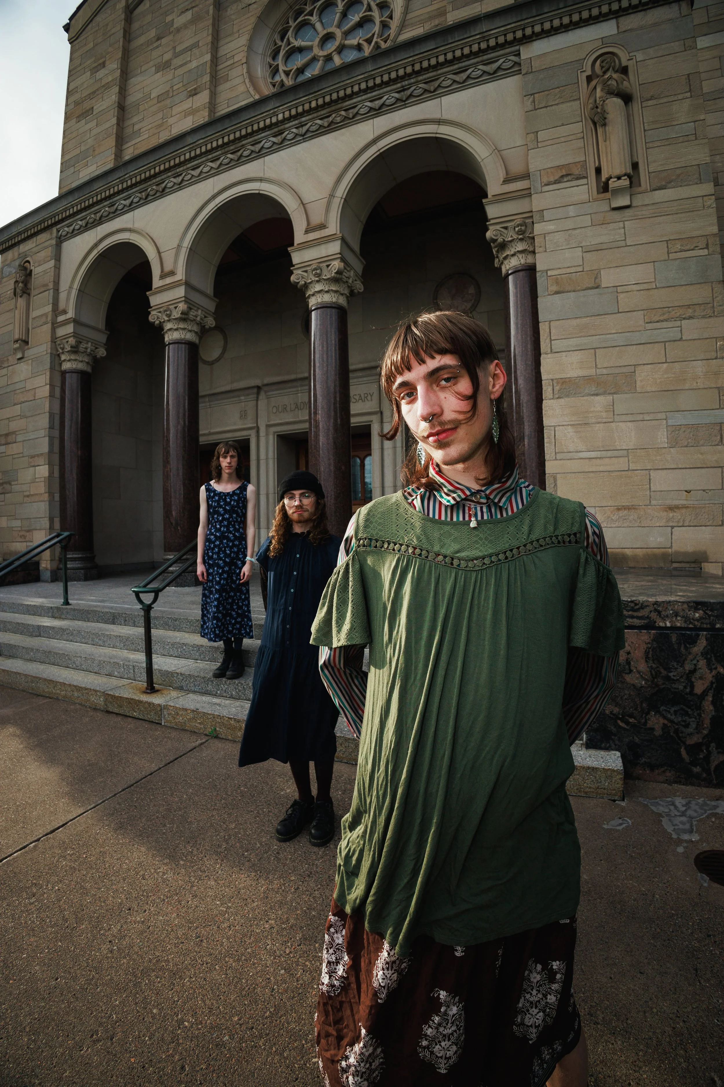 Four young individuals standing on the steps of a stone building with columns and decorative details. The person in the foreground has short brown hair, a septum piercing, and is wearing a green dress over a striped shirt. The others are in the backg