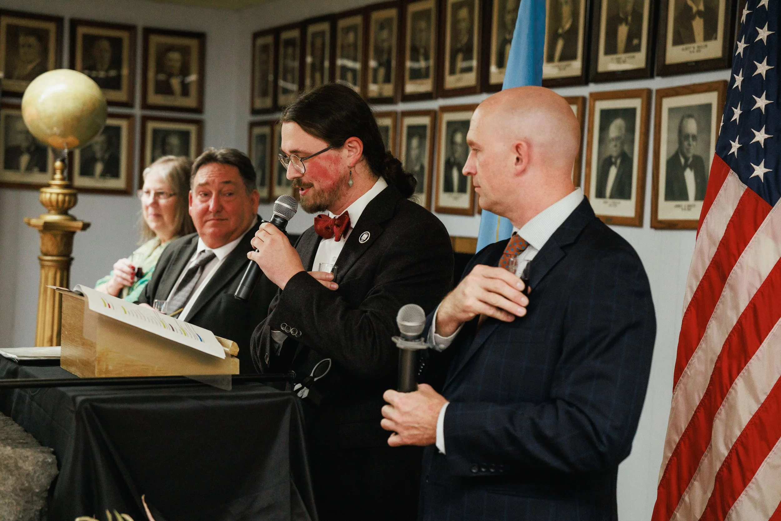 Four people are sitting at a table, with framed photographs on the wall behind them. Two men are holding microphones, and a woman is sitting in the background. The man with glasses and a beard is speaking into the microphone, while the other man is h