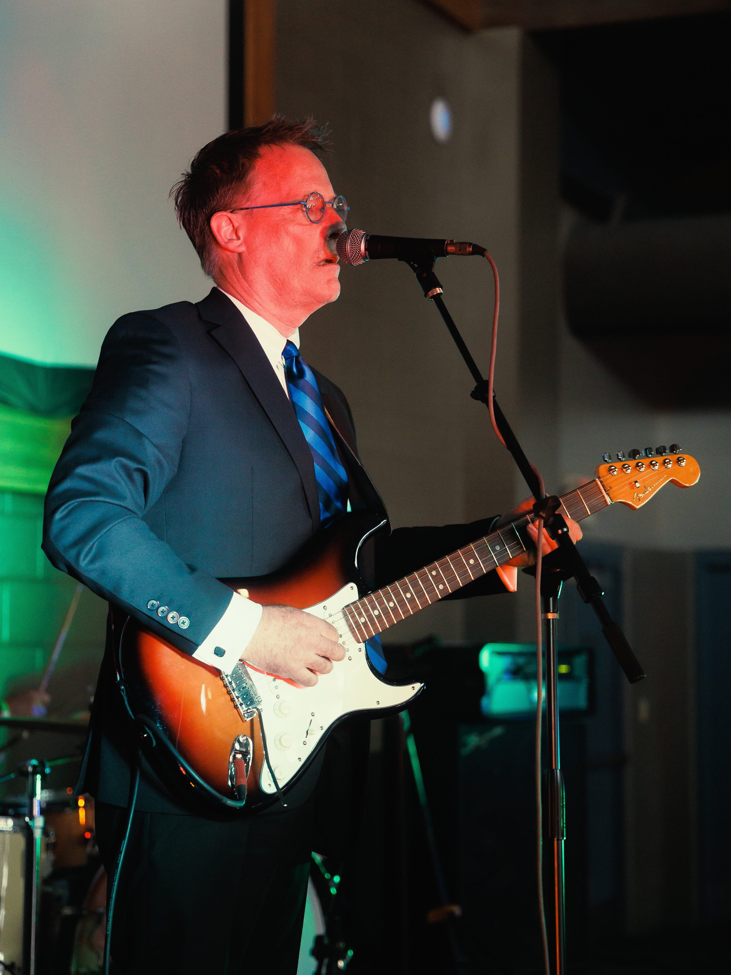 A man in a suit and glasses singing into a microphone while playing an electric guitar during a performance.