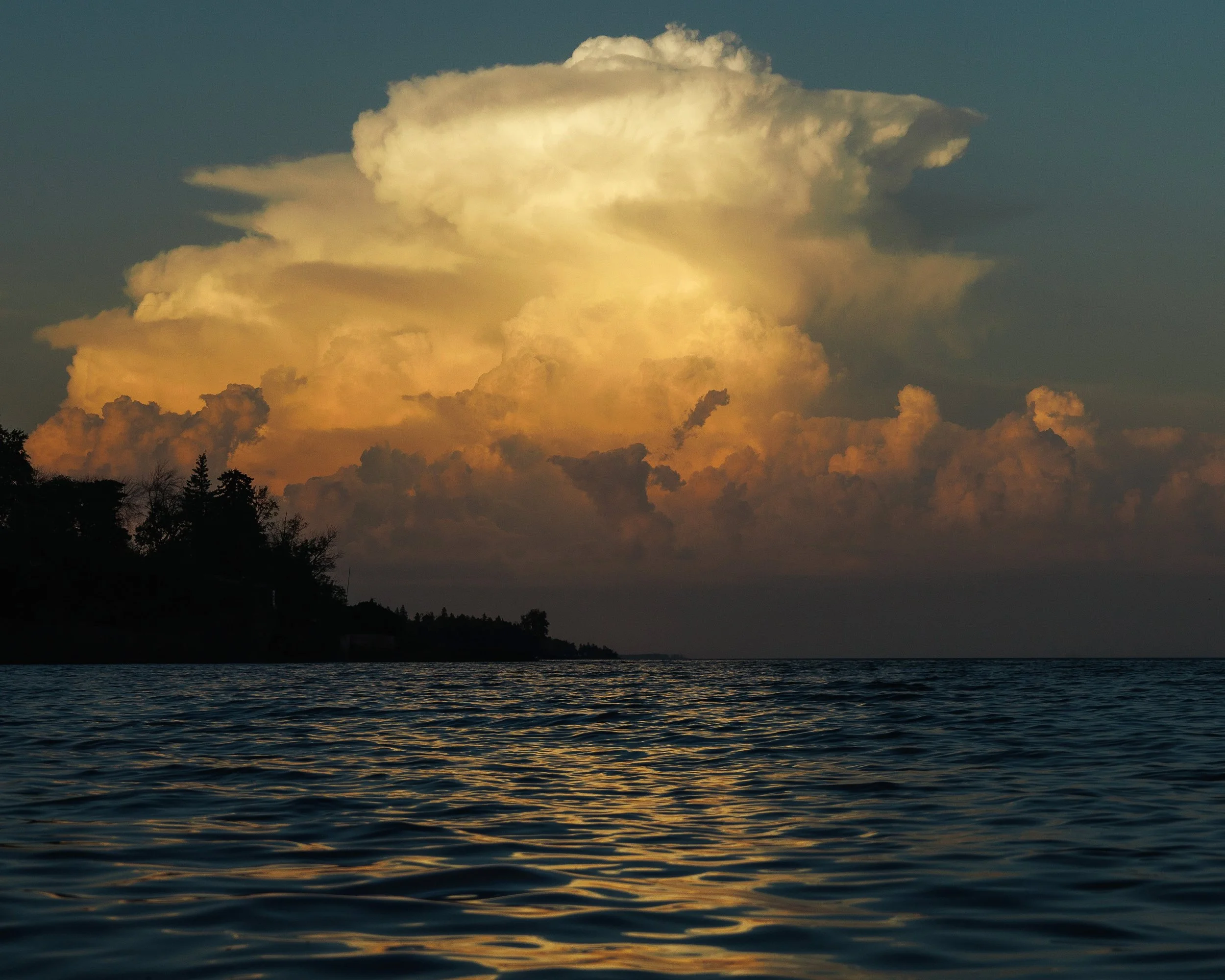 A large, billowy cloud formation illuminated by the setting sun over a body of water, with a dark silhouette of trees along the shoreline in the foreground.