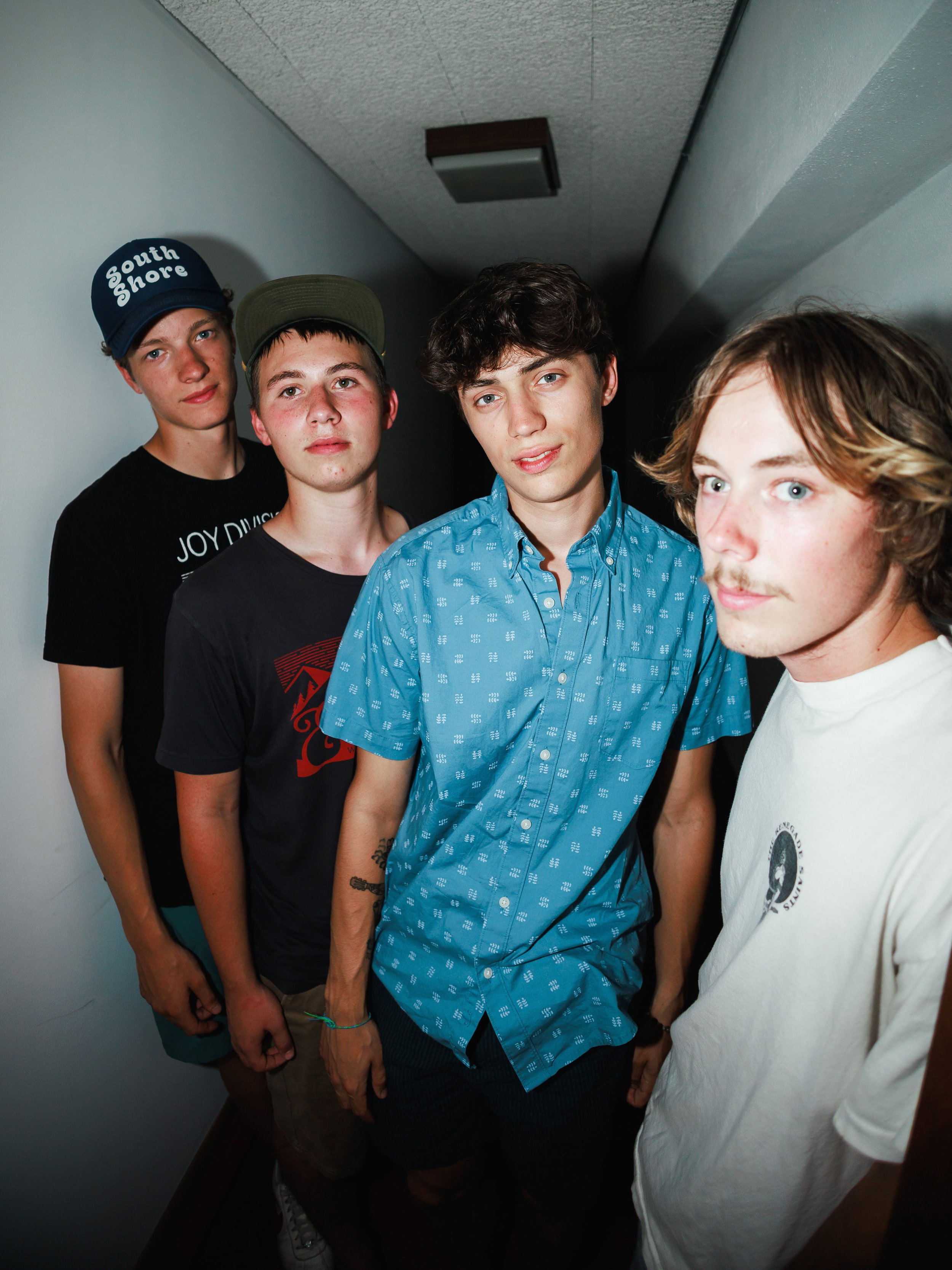 A group of four young men standing in a narrow hallway. They are facing the camera, with the first man on the right partially visible, the second man in the center wearing a blue patterned shirt, the third man on the left wearing a black t-shirt and 