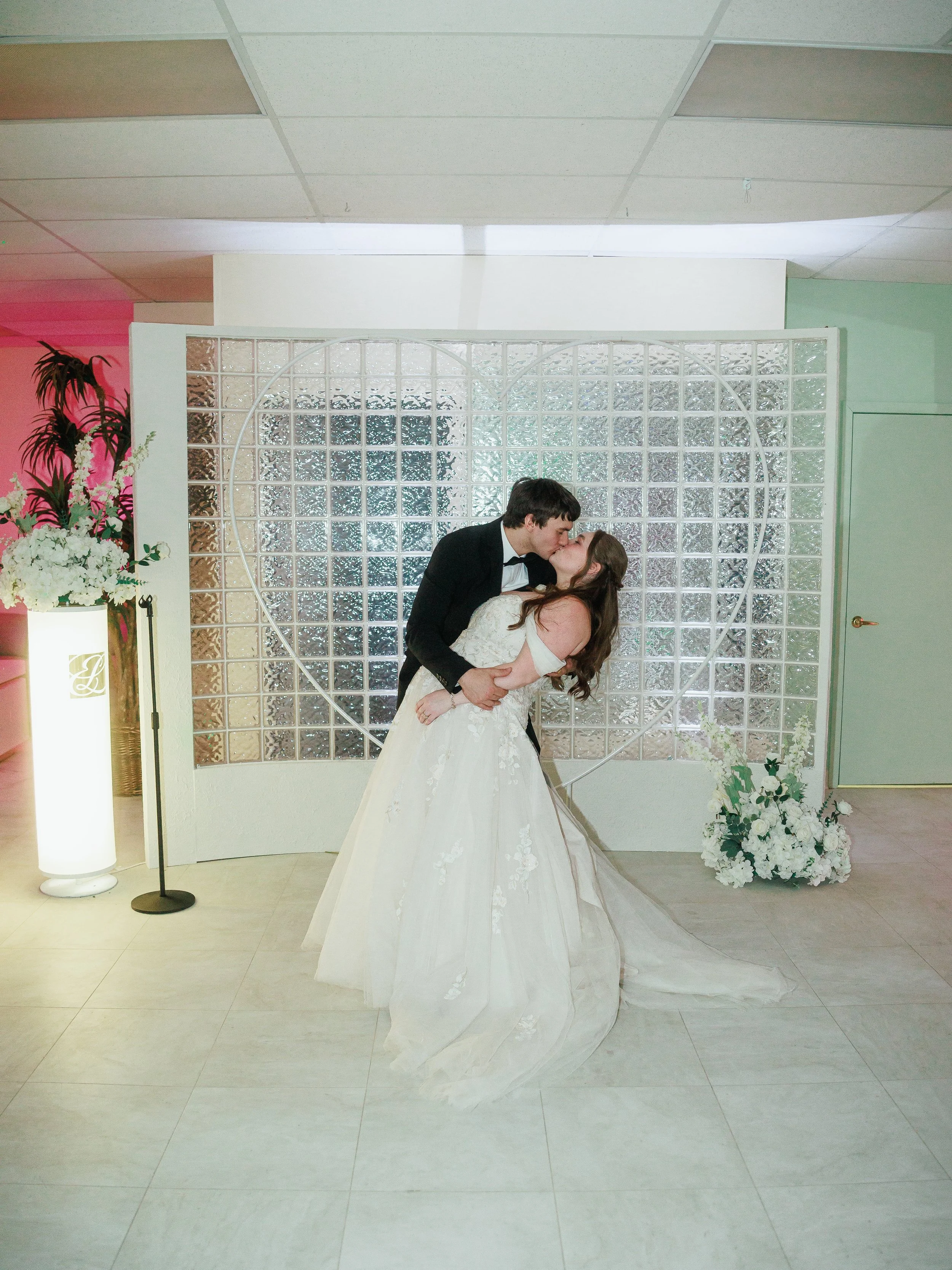 A newlywed couple sharing a kiss on their wedding day, standing in front of a decorative heart-shaped backdrop with floral arrangements.