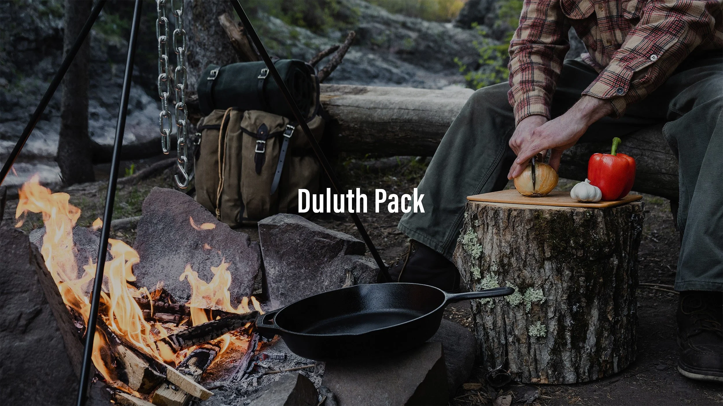 A person preparing food near a campfire in an outdoor forest setting. The person is chopping a garlic bulb on a tree stump, with a red bell pepper and garlic on a cutting board. Camping gear and backpacks are in the background.
