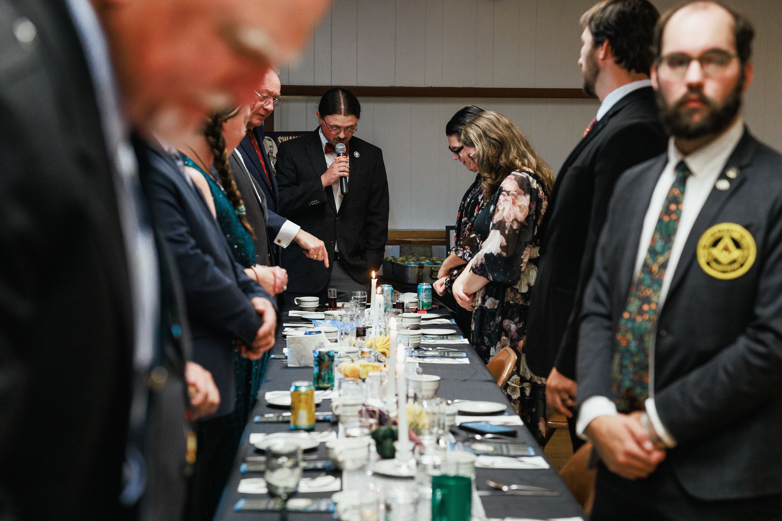 People standing around a long dining table with candles, dishes, and drinks, bowing their heads in prayer at a formal dinner event.