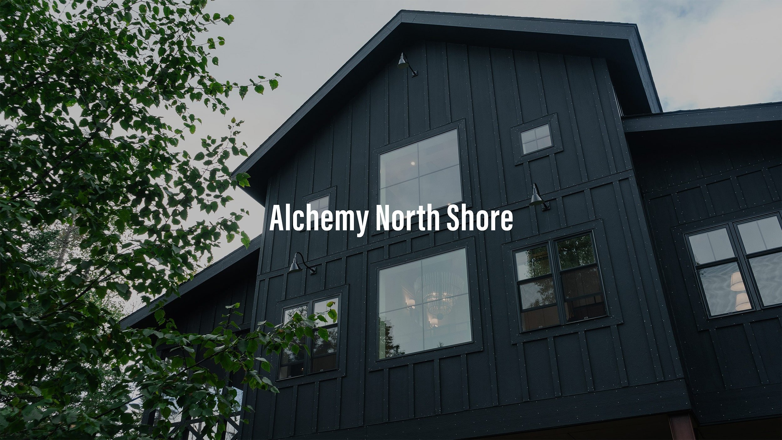 A black modern house with large windows and a steep gabled roof, partially obscured by a tree with green leaves, with the text "Alchemy North Shore" overlaid.