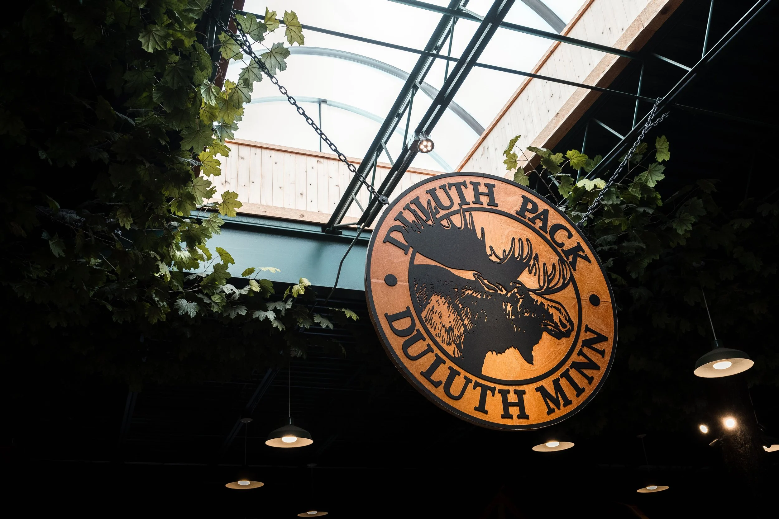 A circular sign hanging from a ceiling, displaying a moose and the words "Duluth Pack" and "Duluth, Minn." The sign is surrounded by green foliage with a skylight and metal beams above.