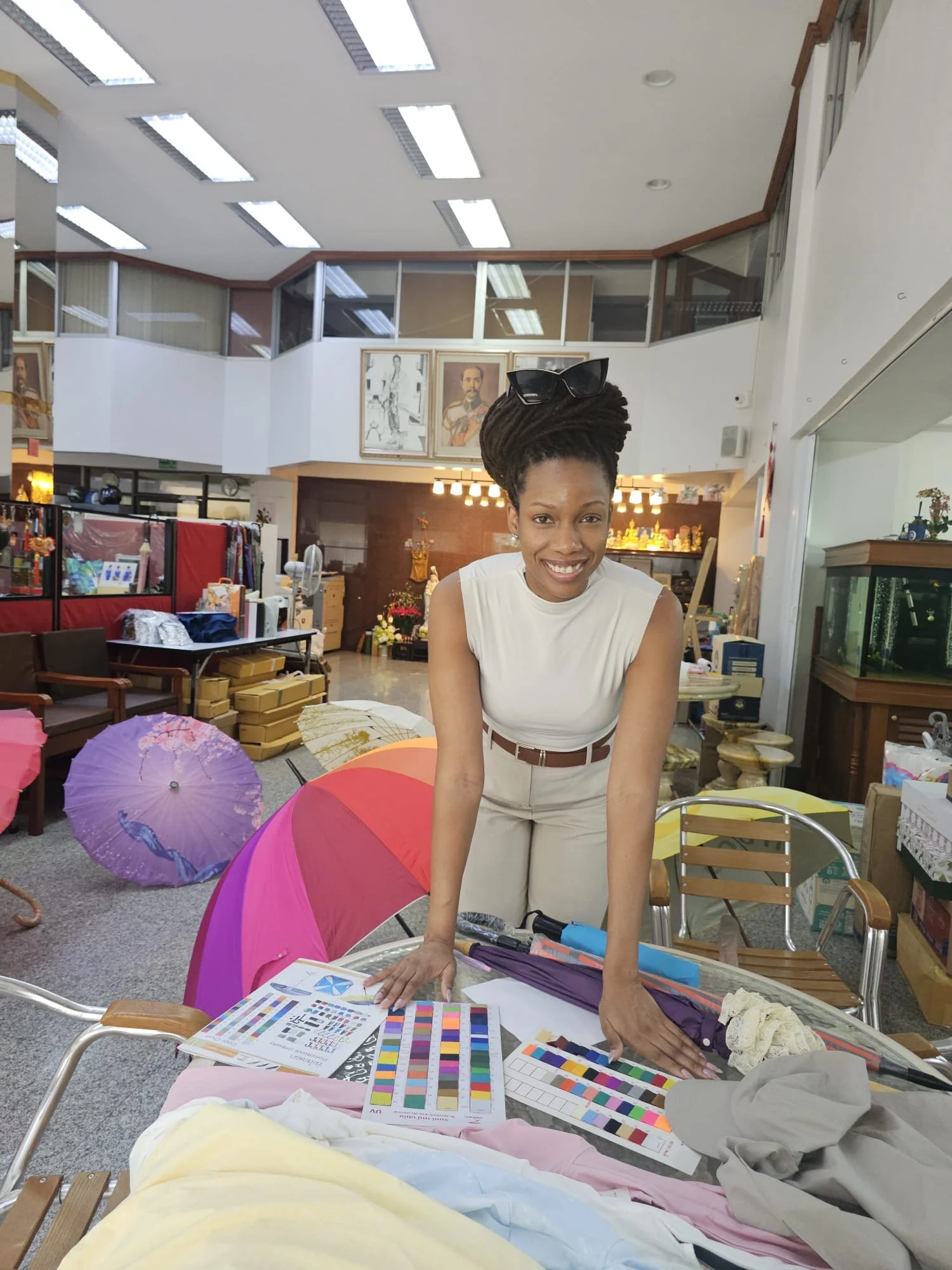 A woman with dreadlocks and sunglasses on her head, smiling, standing behind a table with fabric samples and color charts in a fabric or craft store.