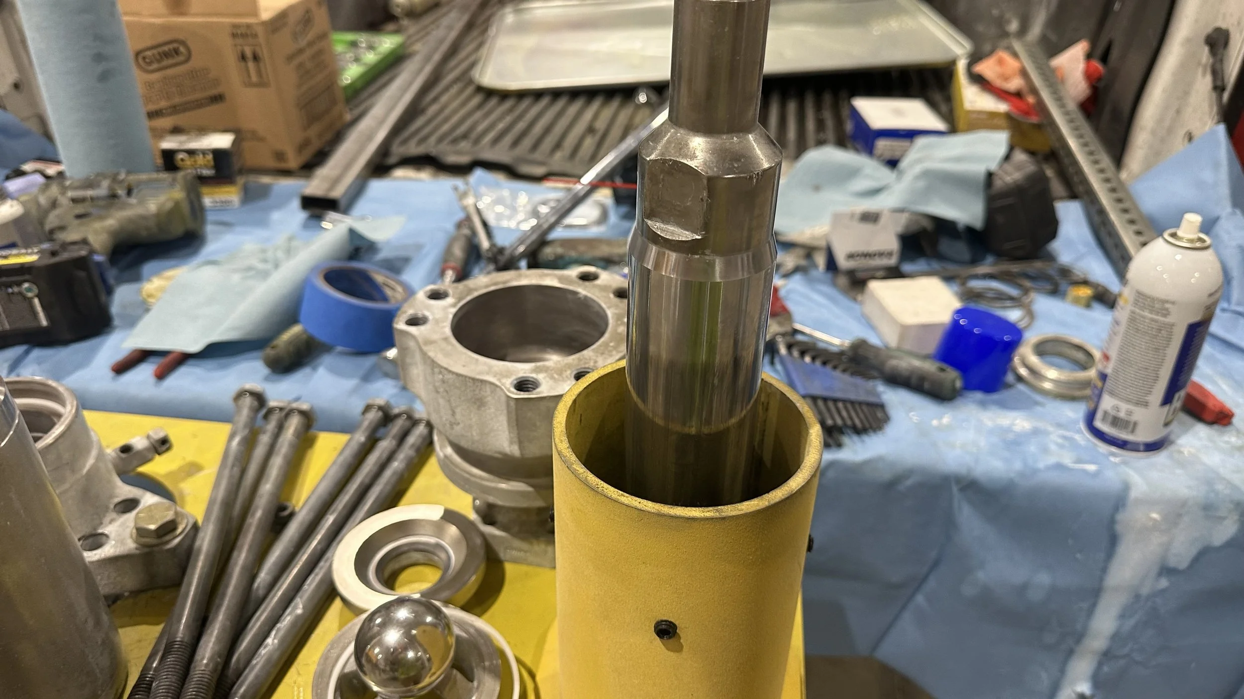 Tools and parts on a workbench, including a metal shaft, various bolts, washers, a bearing, and blue painter's tape, with a yellow container and a cluttered background of boxes, lubricants, and tools.