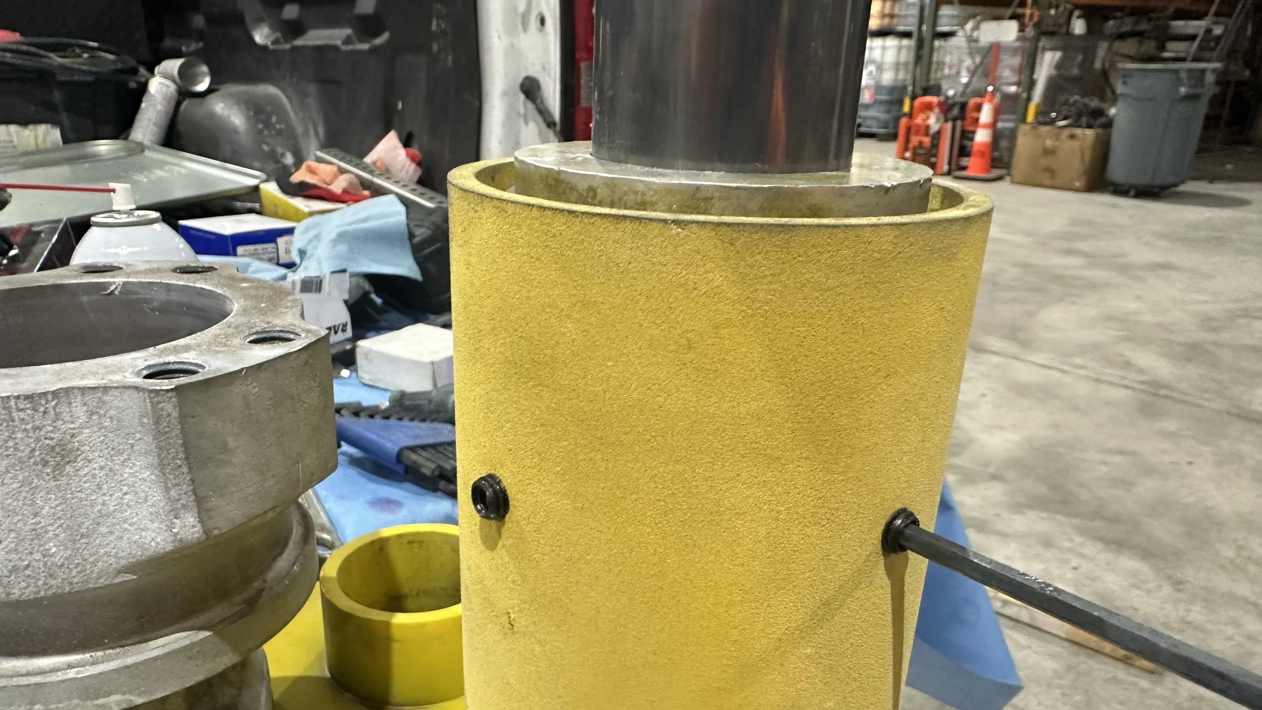 A yellow foam-covered cylindrical component with a metal rod inserted through a hole, in a workshop with various tools and parts on a blue work surface.