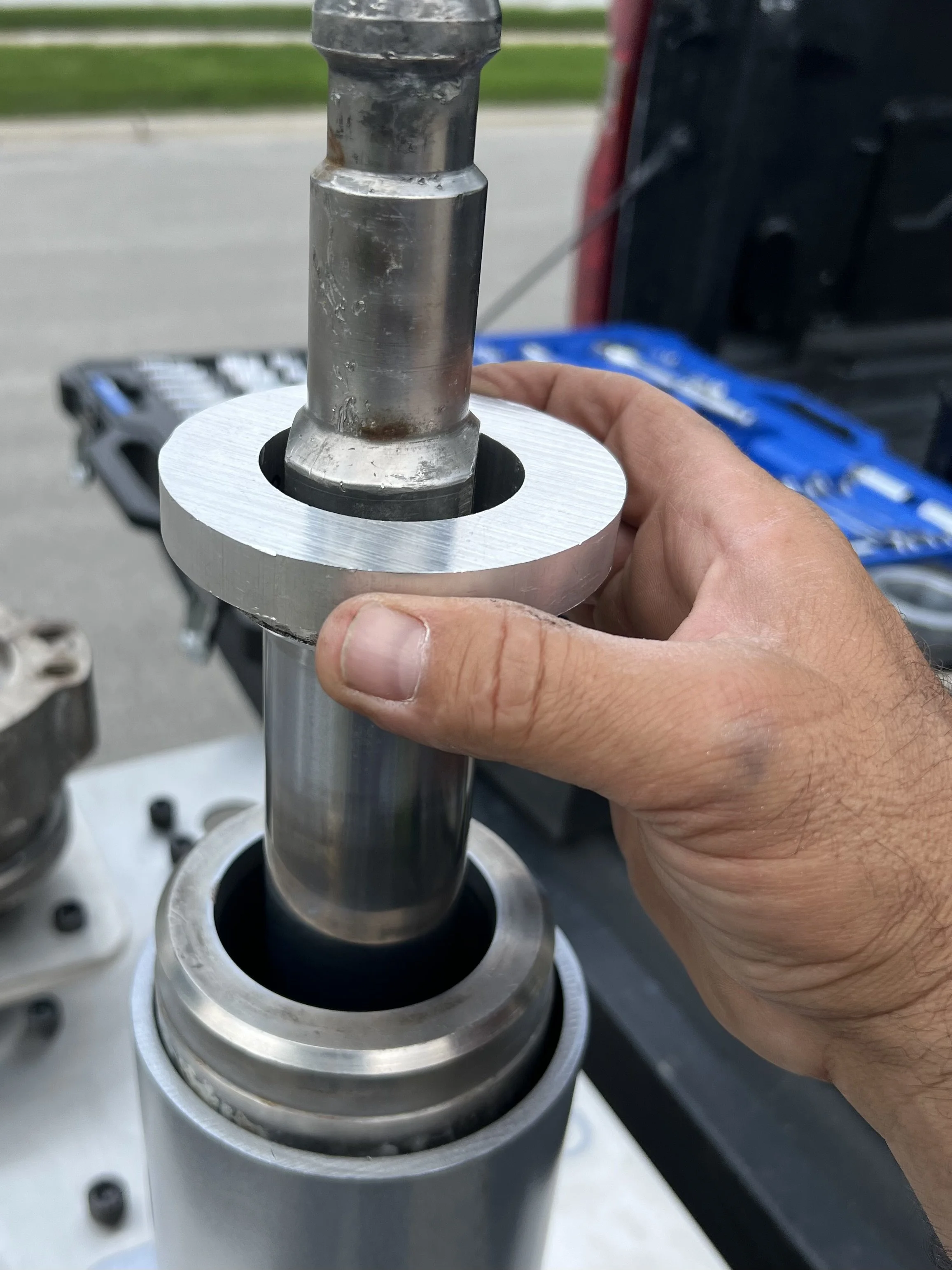 A person holding a metal component with a black rubber seal at the top, being installed into a cylindrical mechanical part outdoors.