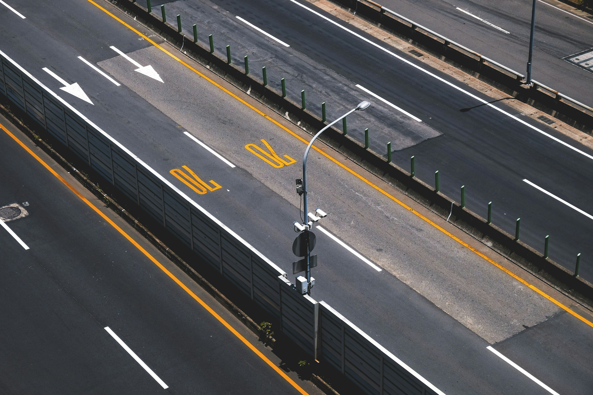 An empty multi-lane road with multiple white arrows and speed limit markings, divided by a physical barrier and a pole with security or traffic cameras.
