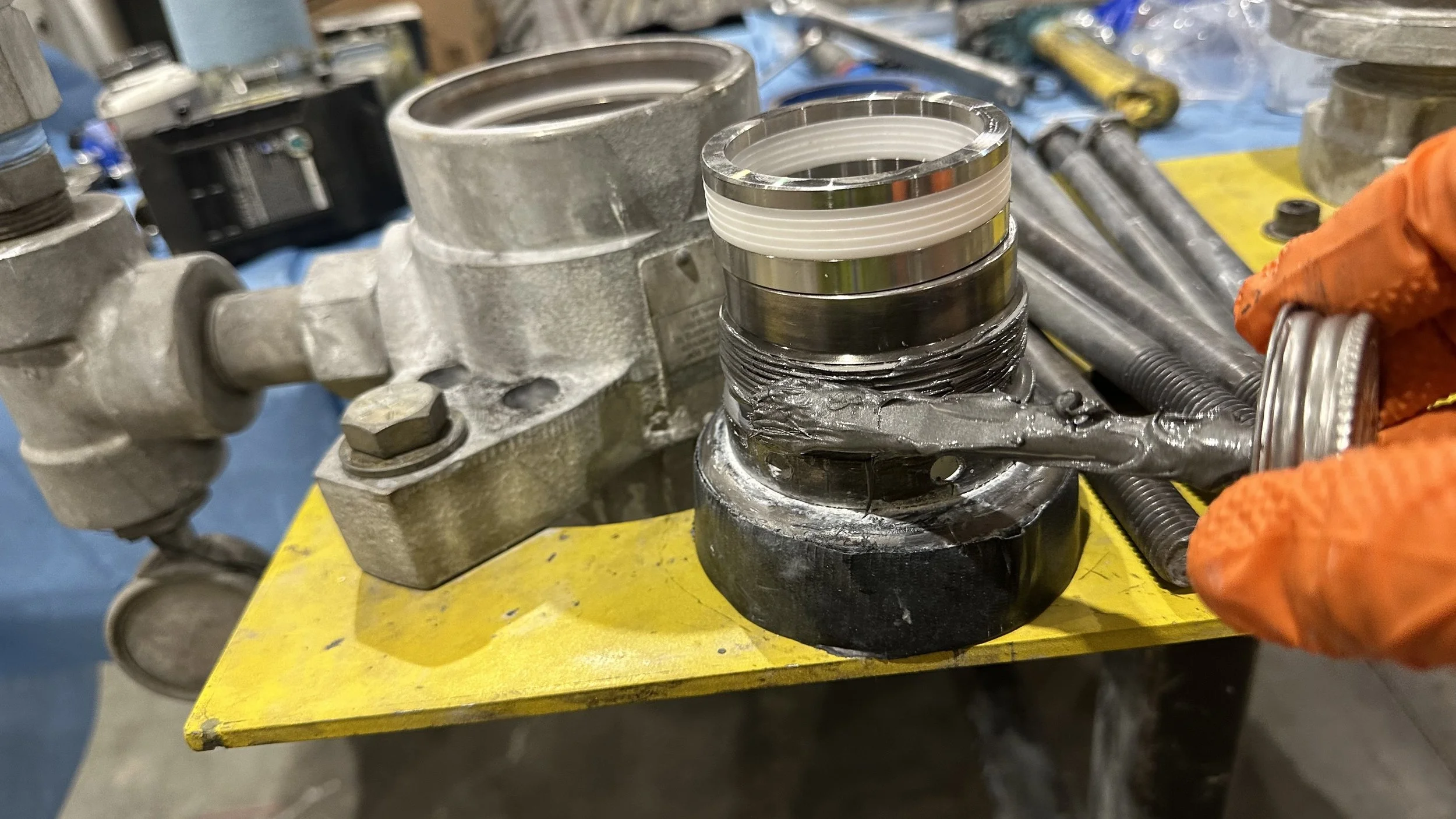 A close-up of a mechanic's hand wearing an orange glove, holding a new piston with a metal crown and white rings, positioned over a black base on a yellow workbench. Surrounding tools include large metal components and threaded rods.