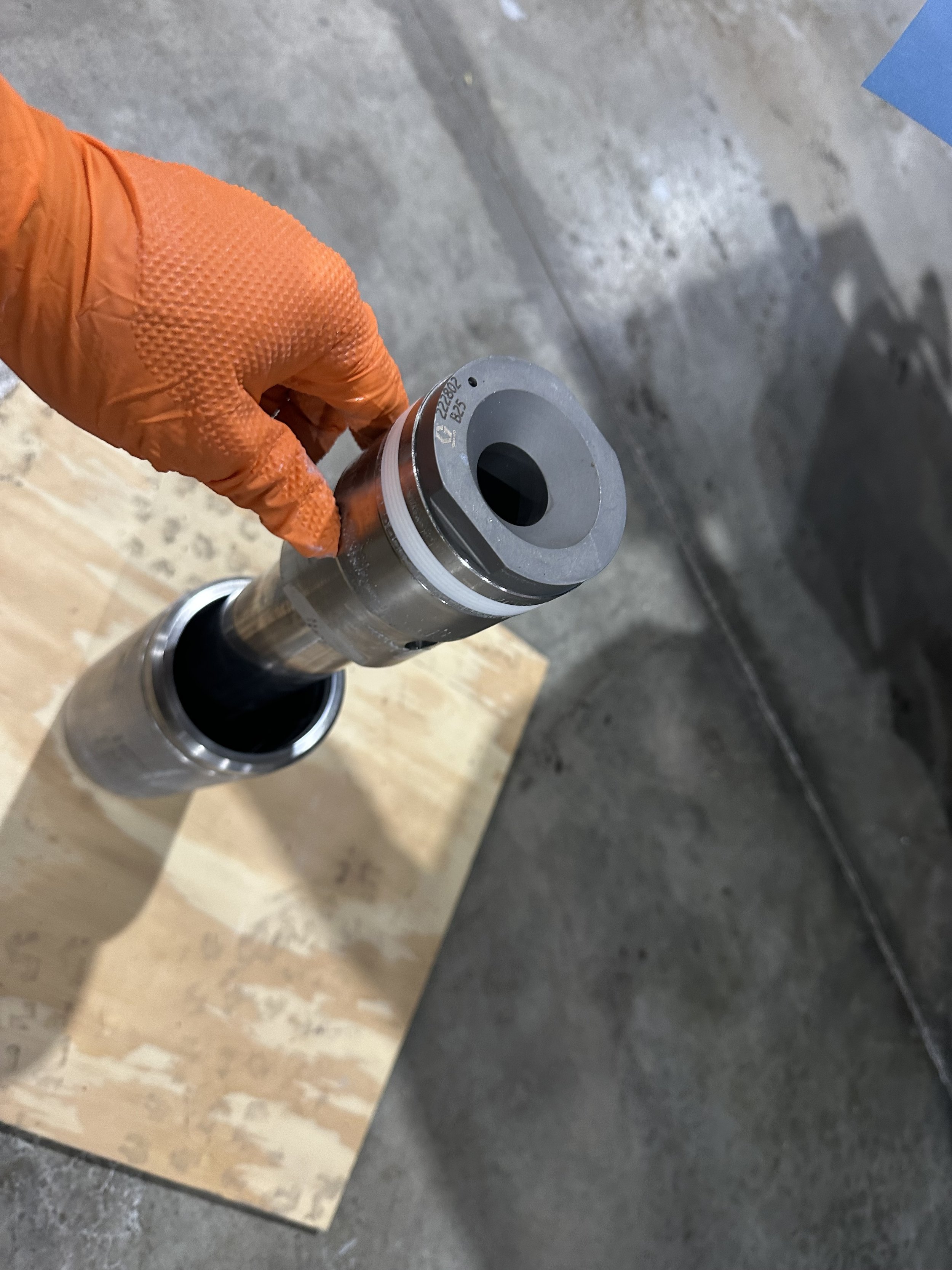 Person wearing an orange glove holding a metal hydraulic or mechanical component with a black interior, standing on a wooden block on a concrete floor.