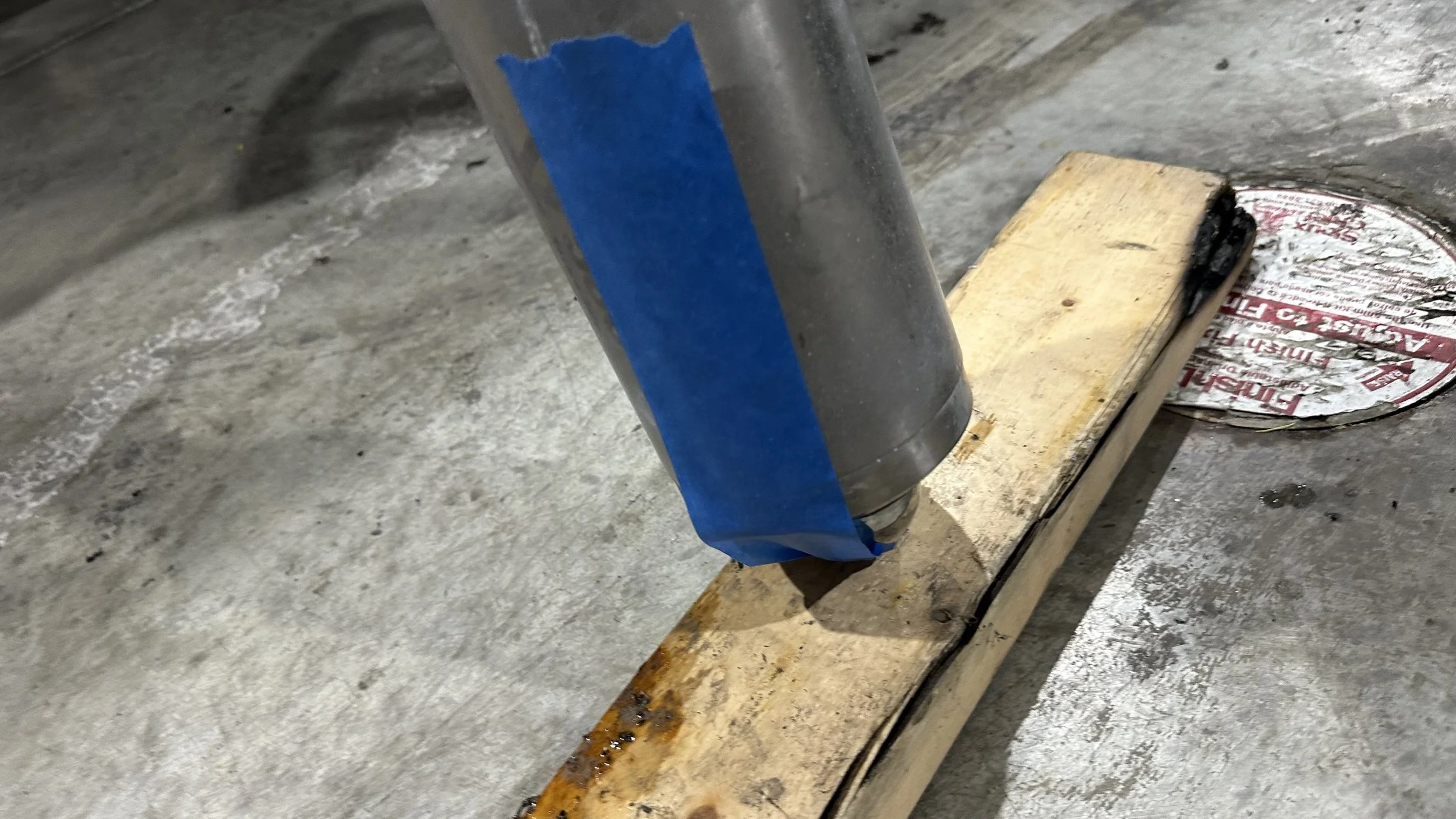 A metal pipe taped with blue painter's tape is mounted upside down on a wooden plank placed on a concrete floor. Near the pipe, there is a round metal lid with red and white markings and some black substance on top.
