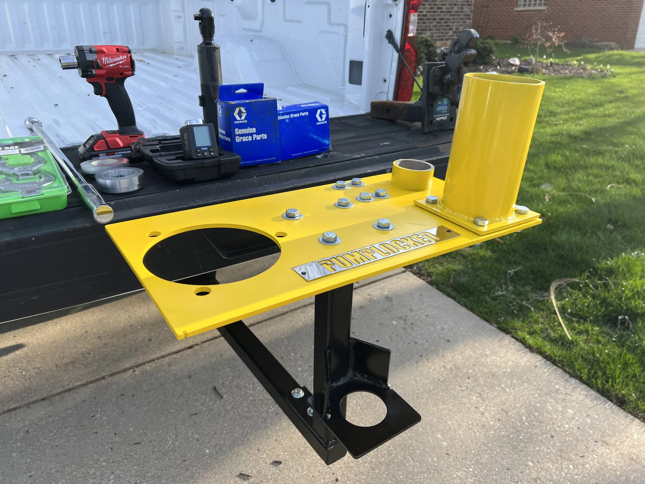 A yellow metal fixture labeled 'PUMPLOCKED' with bolts and cylindrical tubes, mounted on a black metal stand, is placed on a Pickup truck hitch with tools and parts spread out on a black lift gate in the background.