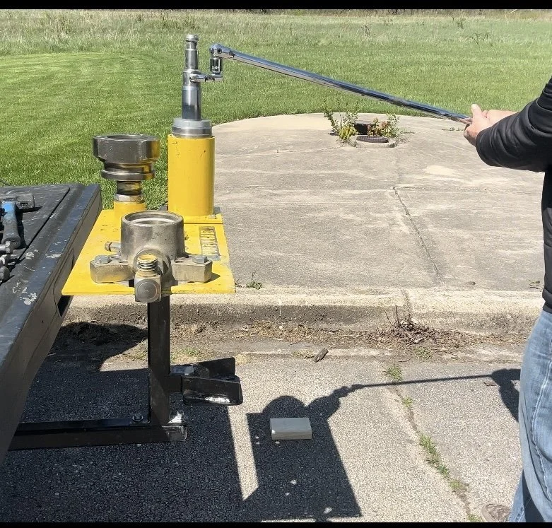 A person appears to be operating a hydraulic press or testing machine outside, with a yellow base and metallic components, on a concrete surface, with grass in the background.