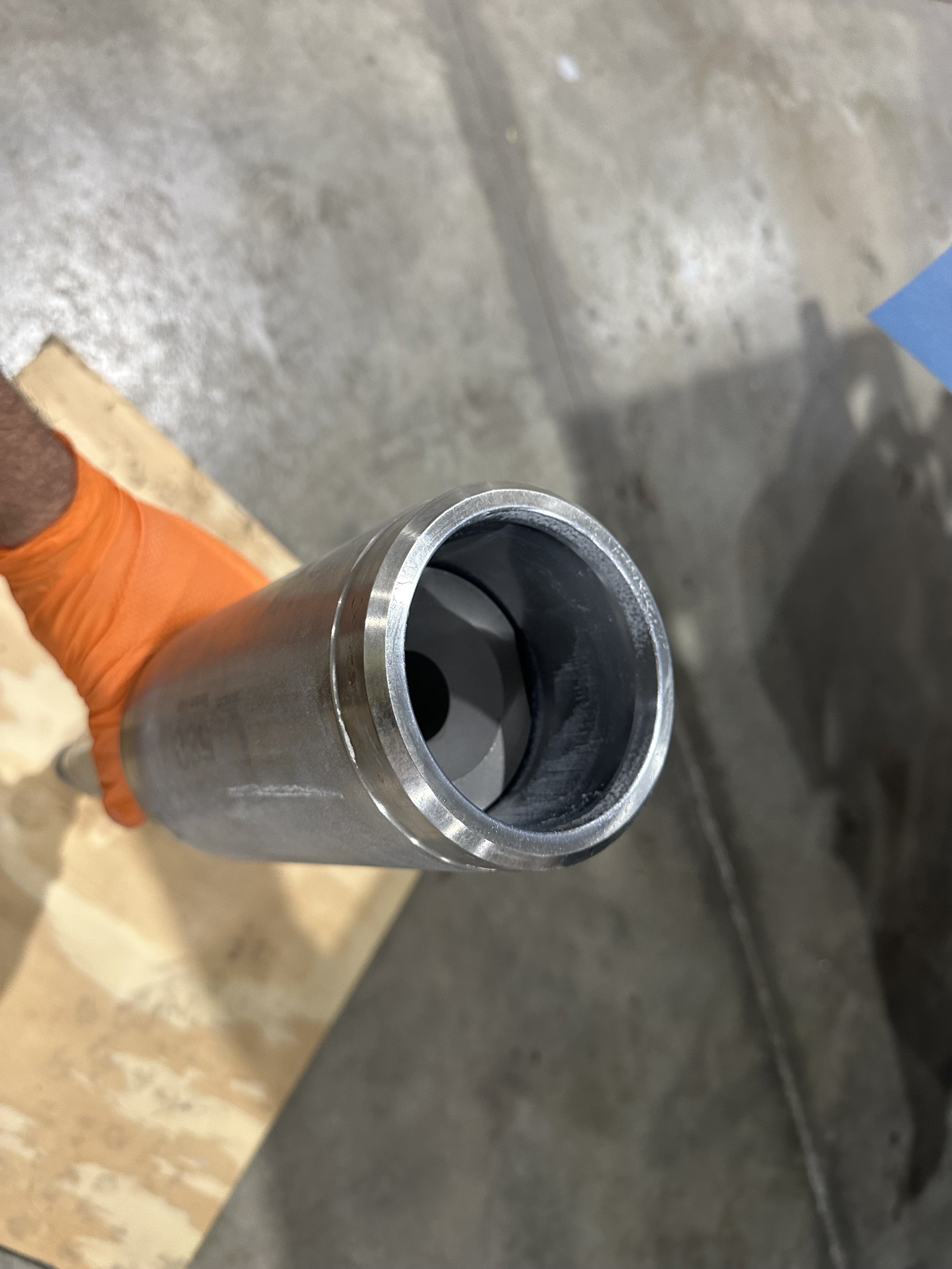 Close-up of a Graco cylinder with a piston metal pipe being held by a person wearing an orange glove, with a concrete floor and some wooden and blue elements in the background.