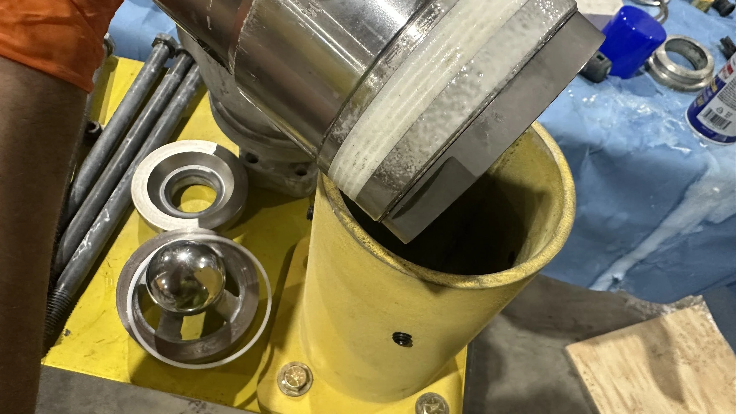 Close-up of a machine with metallic parts, including a large cylindrical component with Graco packings being inserted or removed into a yellow container, with various tools and parts in the background.