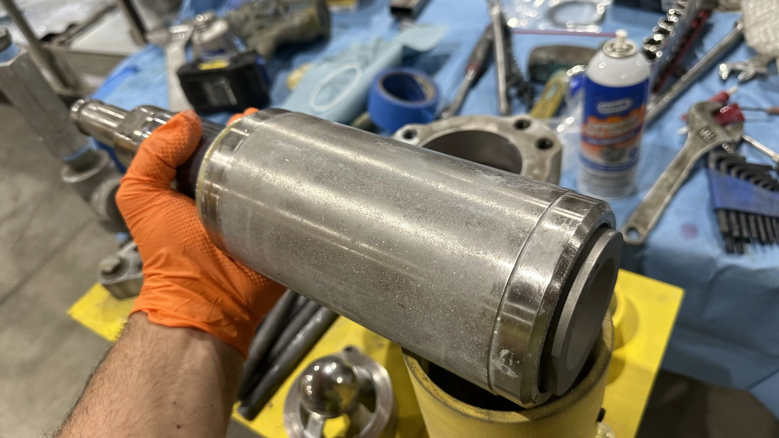 Close-up of a hand in an orange glove holding a metal cylindrical engine part over a blue workbench with various tools and equipment in the background.