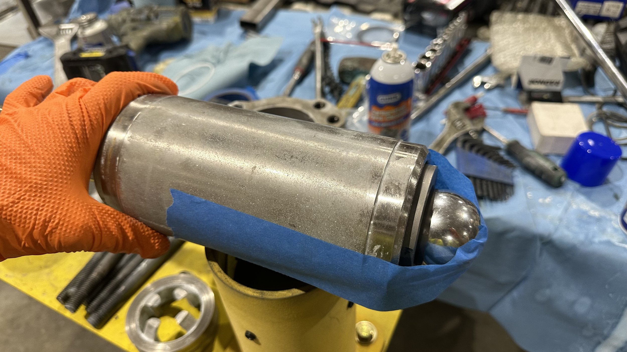 A person wearing orange gloves holding a cylindrical metal object with a rounded end over a yellow container, with a cluttered workbench of tools and supplies in the background.