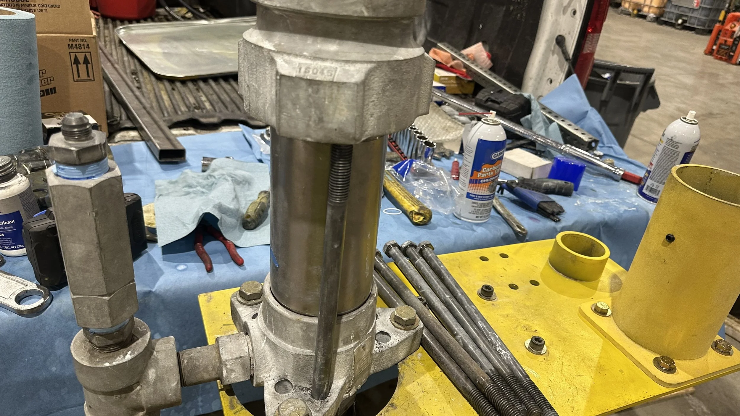 Workshop table with mechanical parts, tools, and supplies, including a yellow testing device and various metal rods and items in the background.