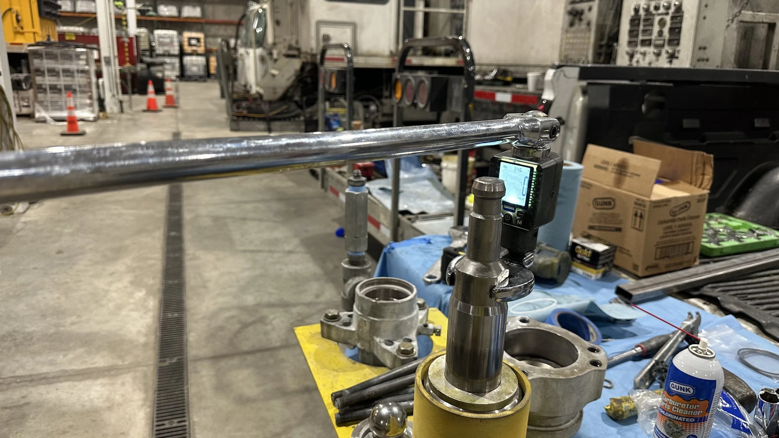 Workshop table with mechanical parts, tools, and a digital setup in a garage or industrial setting, with a truck and various equipment in the background.