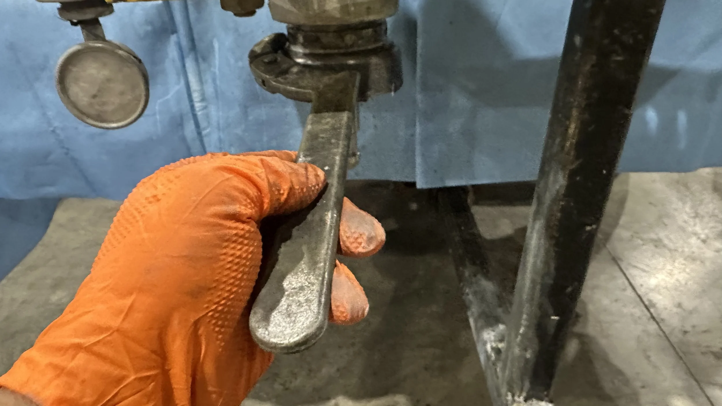 A person wearing an orange textured work glove is holding a metal valve handle near a large blue industrial machine or piece of equipment with pipes.