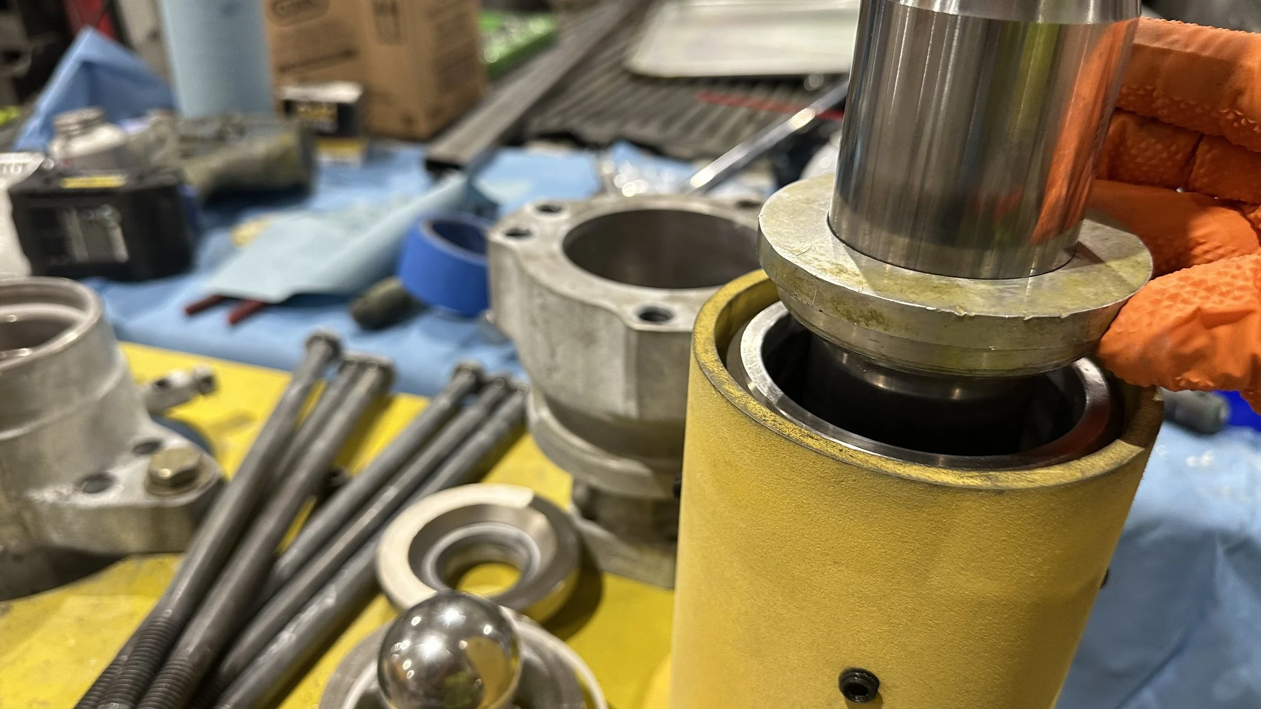 Close-up of mechanical parts and tools on a workbench, showing a person's hand in an orange glove handling a bearing inside a yellow cylindrical component, with various metal components, nuts, and tools scattered around.