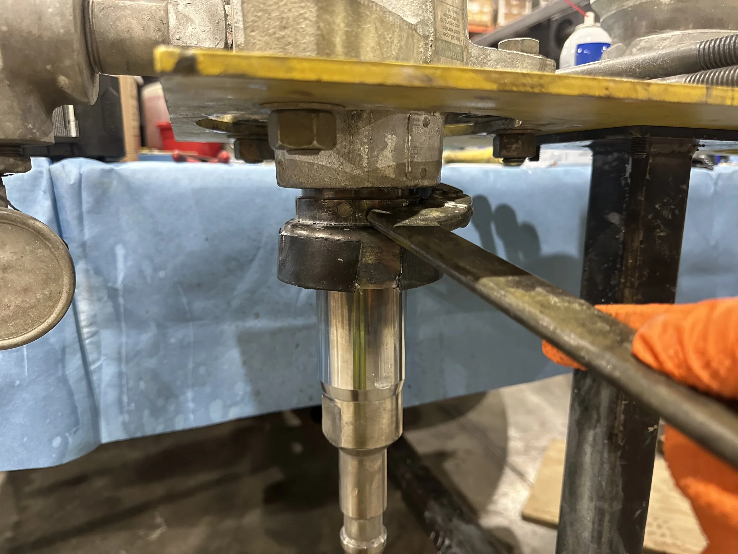 Close-up of industrial machinery with a metal shaft and surrounding components, including a yellow surface and a blue background, possibly in a workshop or factory setting.