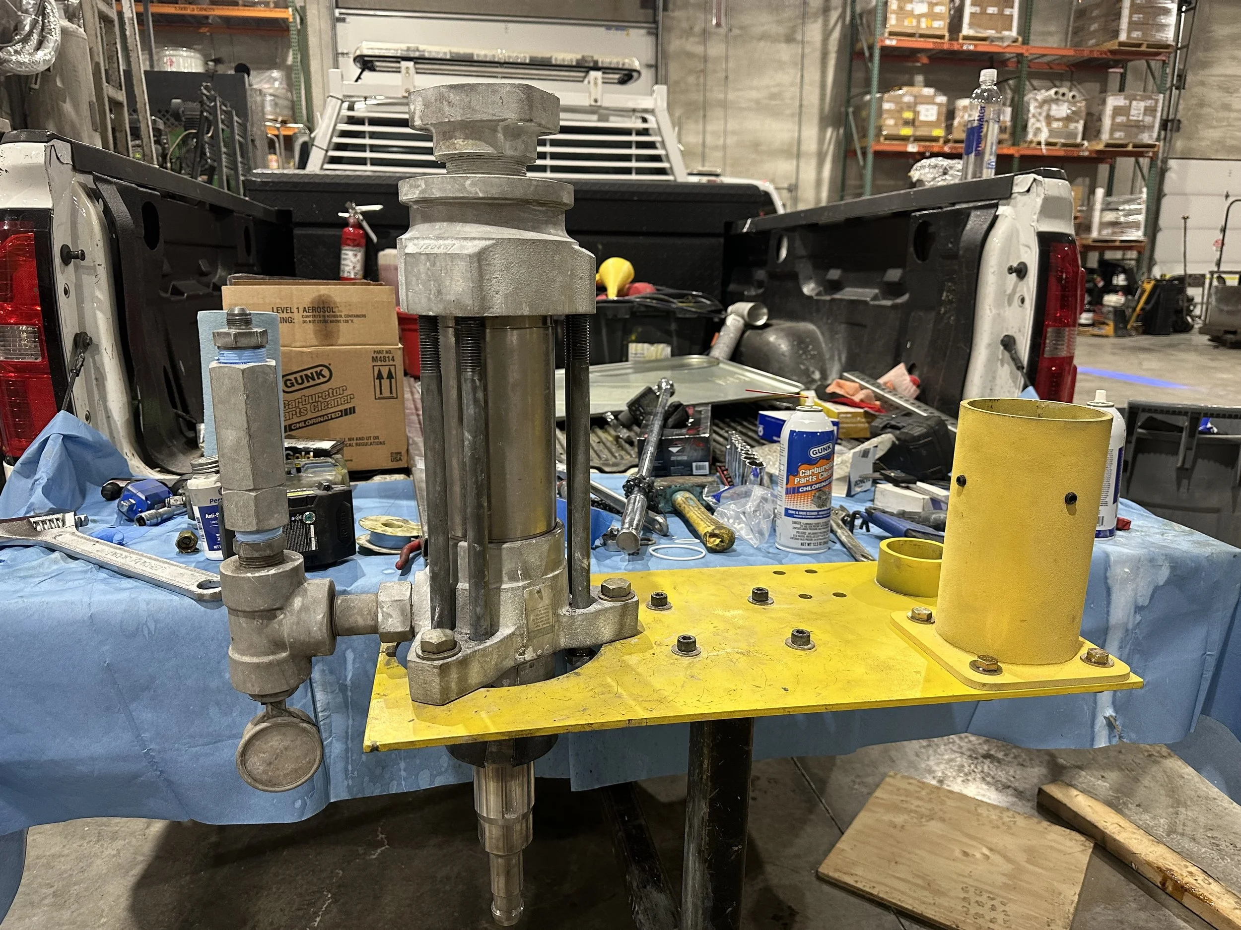 A mechanical device with metal components on a yellow base, placed on a blue cloth-covered table in a workshop setting, with various tools and parts in the background.
