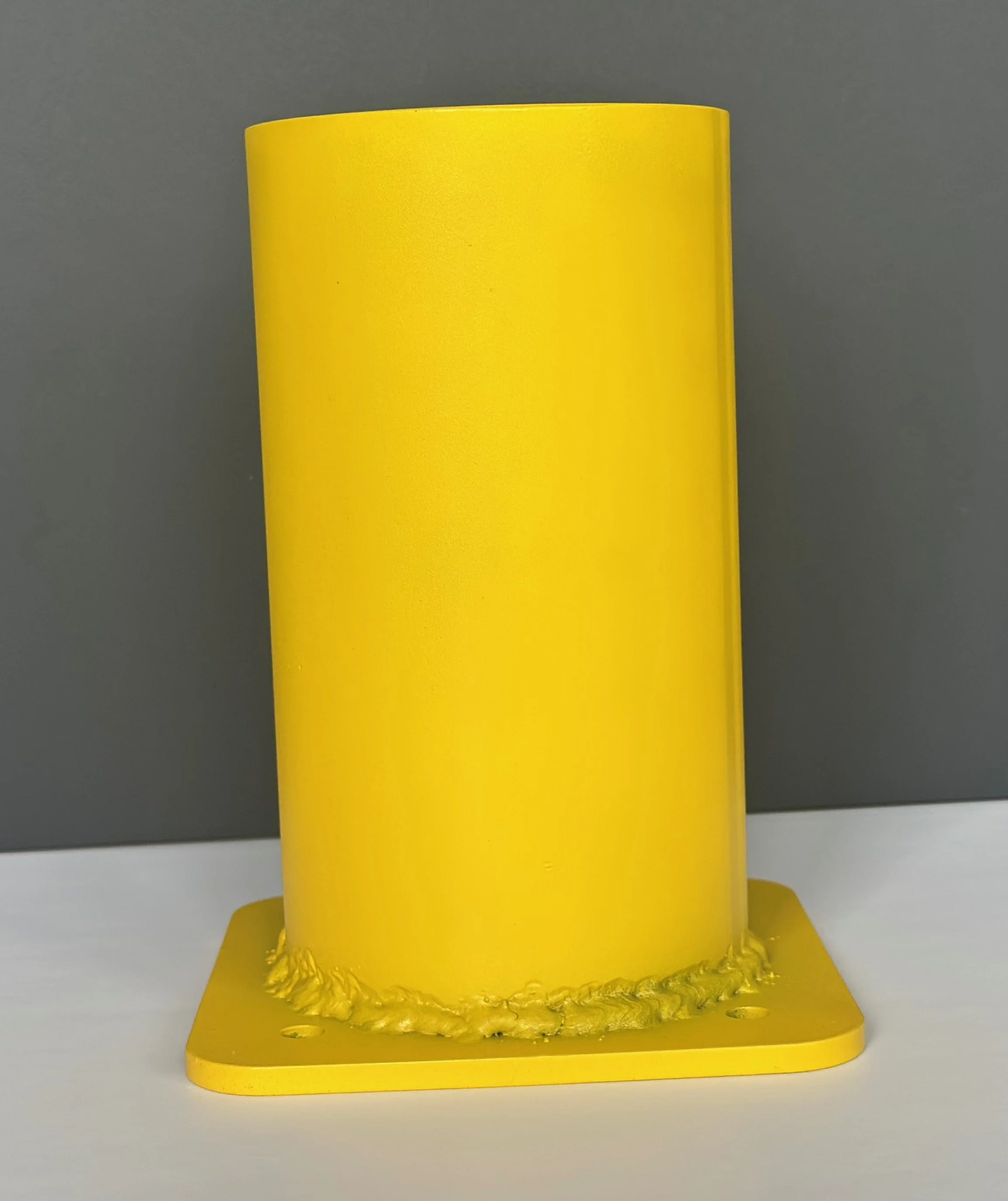 A bright yellow cylindrical object, possibly a container or part of a device, mounted on a yellow square base with visible welding or glue around the bottom edge, against a gray background.