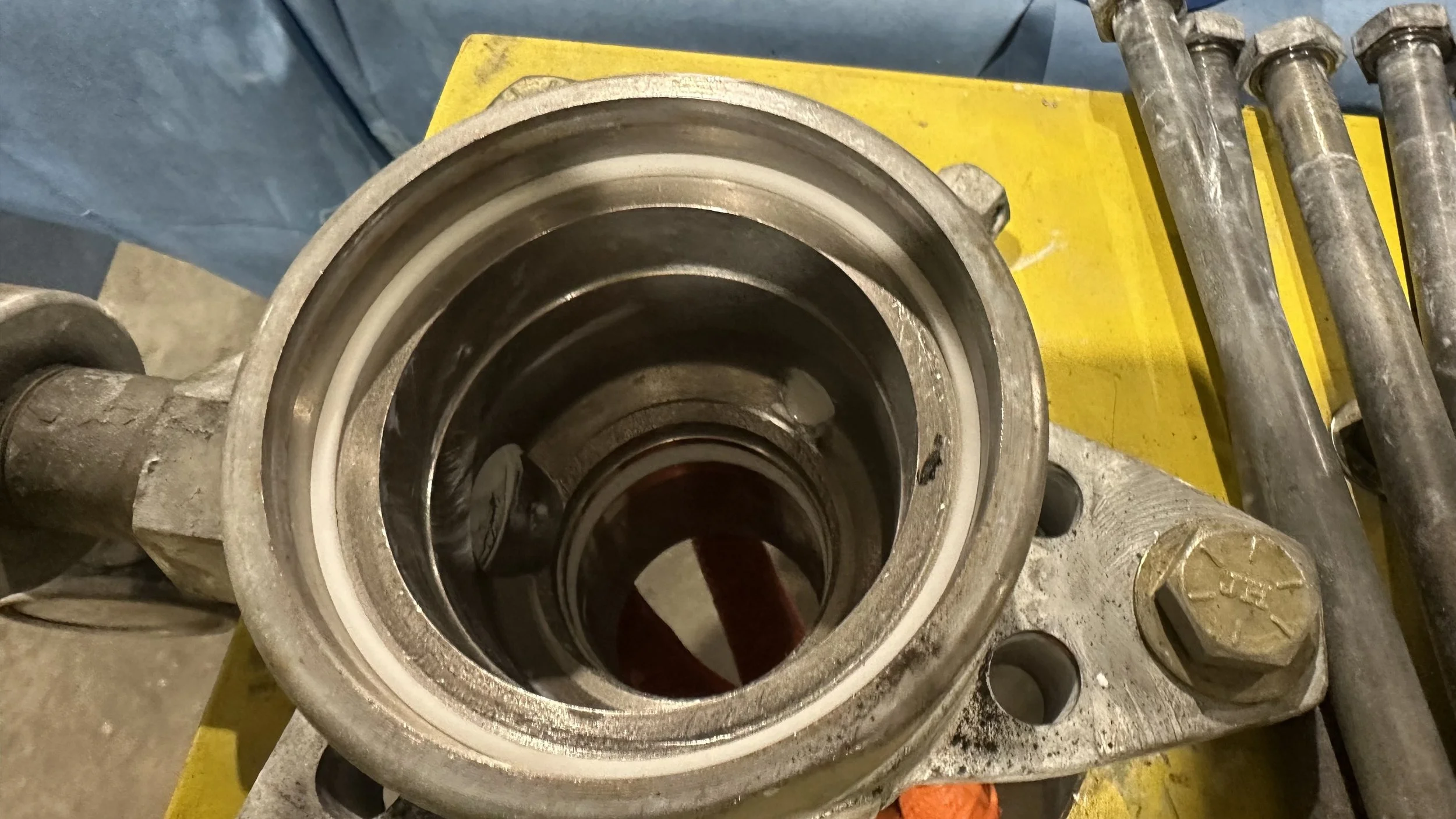 A close-up view of a mechanical component, likely a bearing or part of a machine, with a metal housing, circular opening, and internal rotating parts, placed on a yellow surface with tools around it.