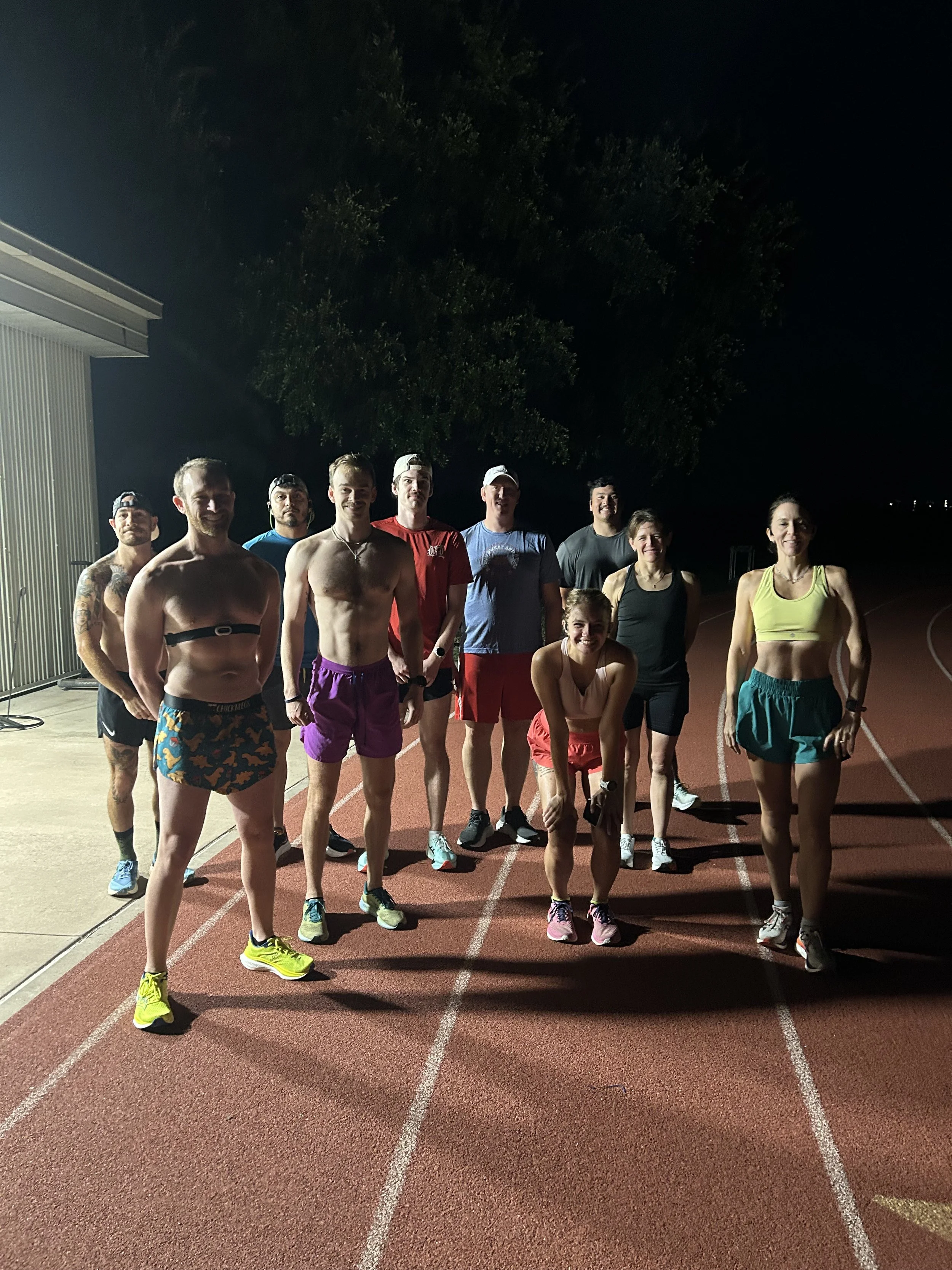 Group of nine runners, both men and women, posing on a running track at night, some in athletic wear and others shirtless, smiling.