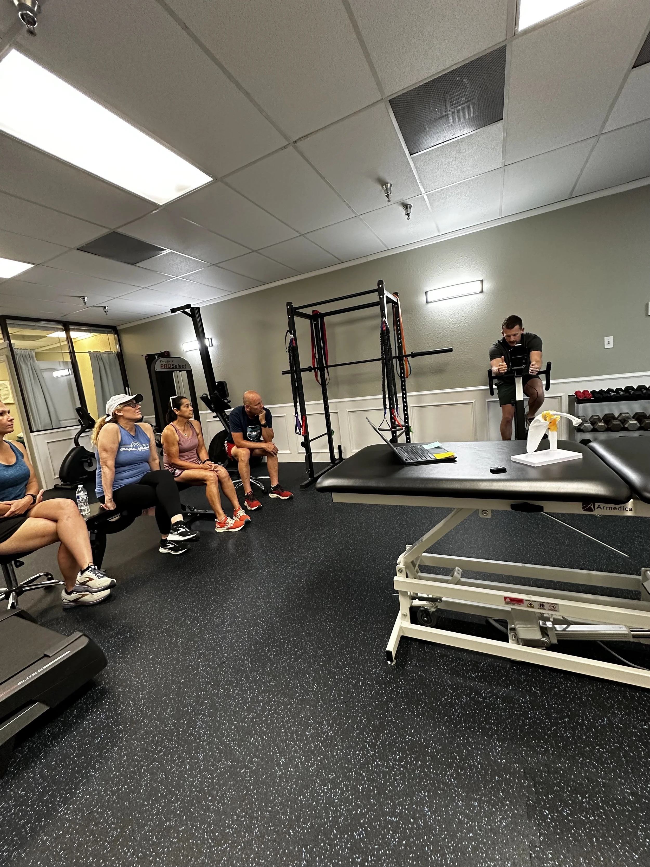 Group of people sitting in a gym, listening to a trainer demonstrating exercise techniques on a stationary bike; gym equipment includes a treadmill, weight rack, and a massage table.