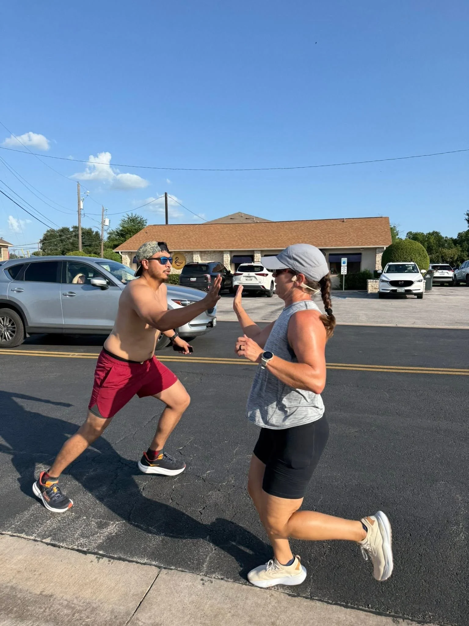 A man and a woman are running outdoors on a sunny day, engaging in a friendly high five as they pass each other on a street with parked cars and buildings in the background.