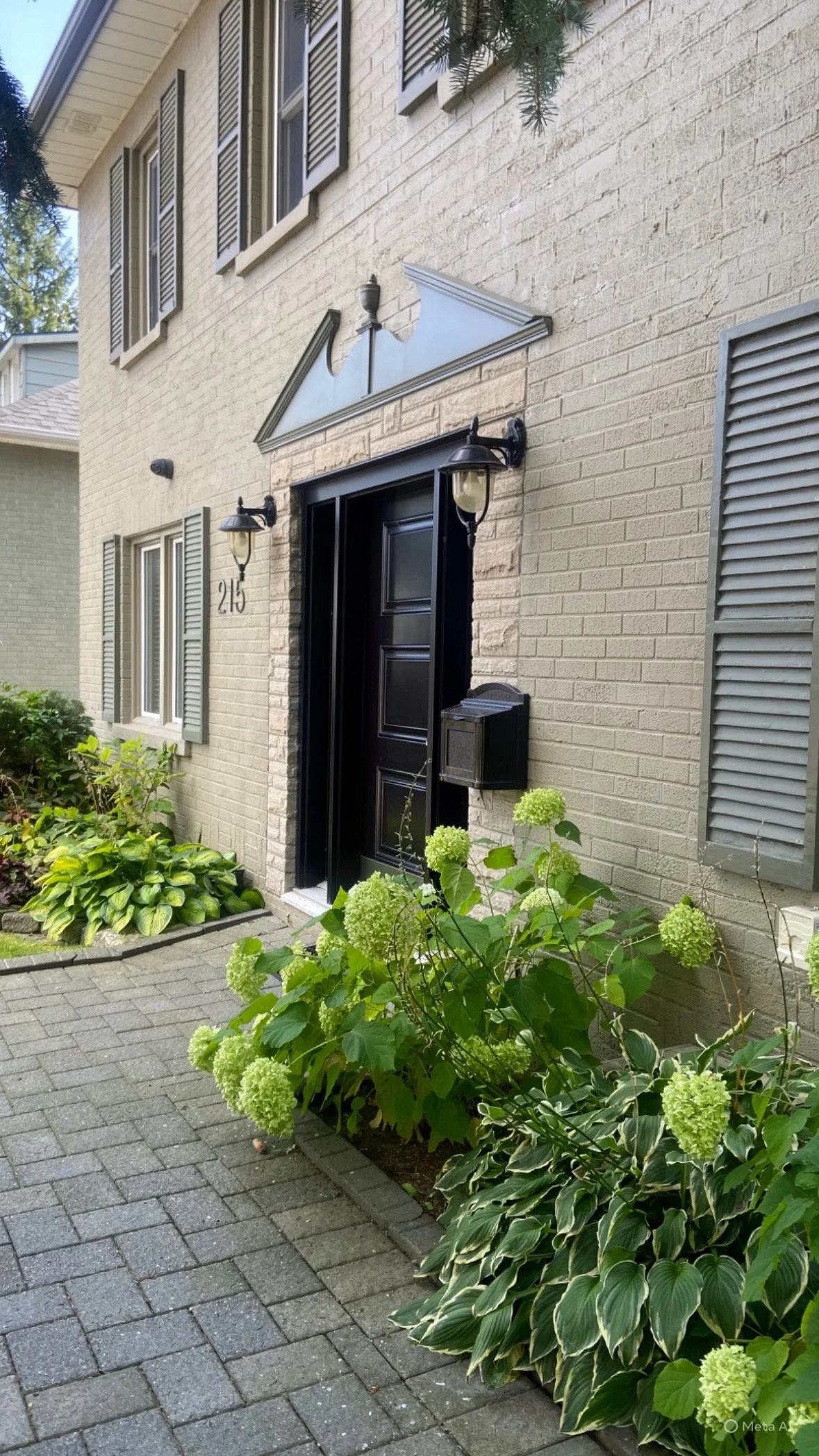 The image shows the front entrance of a house with a black door, a black house mailbox, and two black outdoor lamps on either side of the door. The house is yellow brick and features gray shutters on the windows. In front of the house, there are green bushes with variegated leaves and light green hydrangea flowers, along with a paved brick walkway leading to the entrance.