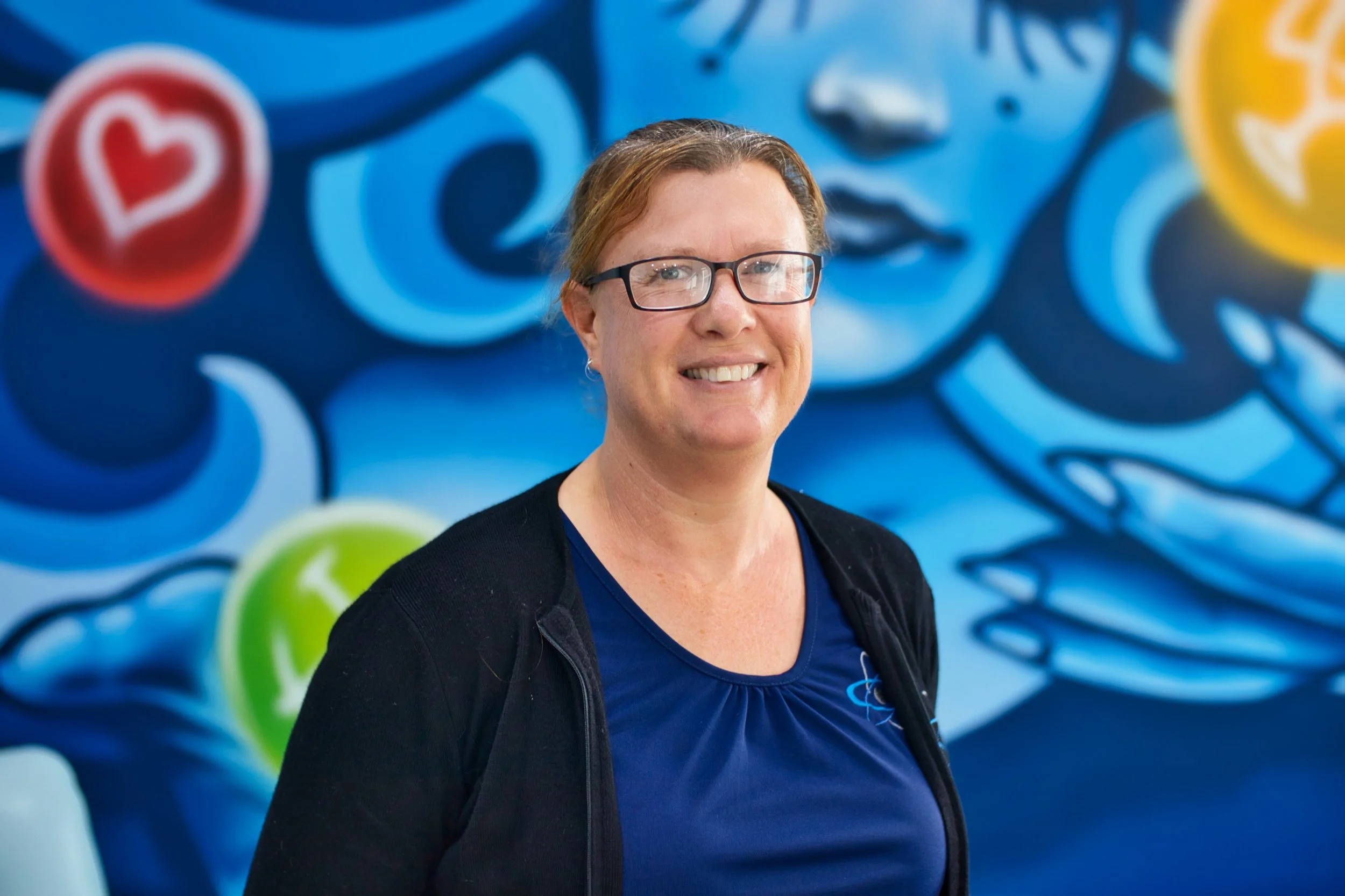 Client Services, Amanda Kenny smiling at camera in front of colourful blue and yellow mural. Located in Bunbury office.