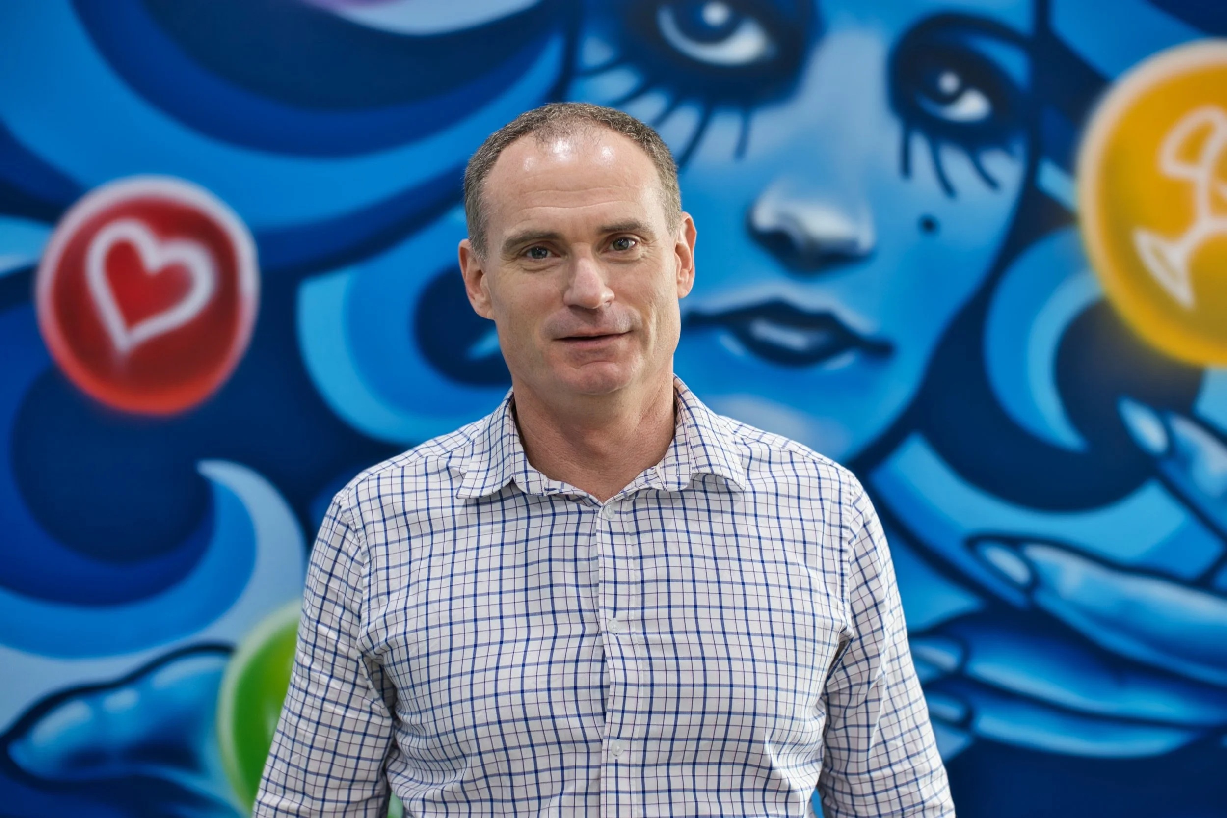 Director Kingsley Smith , smiling at camera in front of colourful blue and yellow mural. Located in Bunbury office.