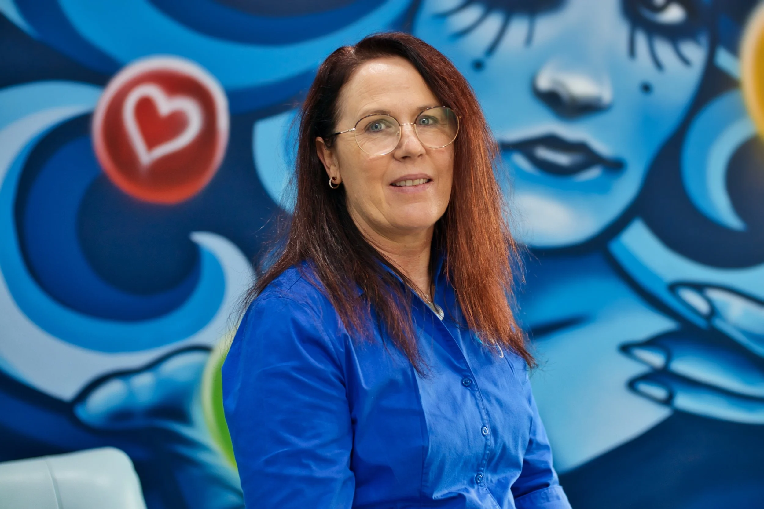 Bookkeeper, Jenny Freeman,  smiling at camera in front of colourful blue and yellow mural. Located in Bunbury office.
