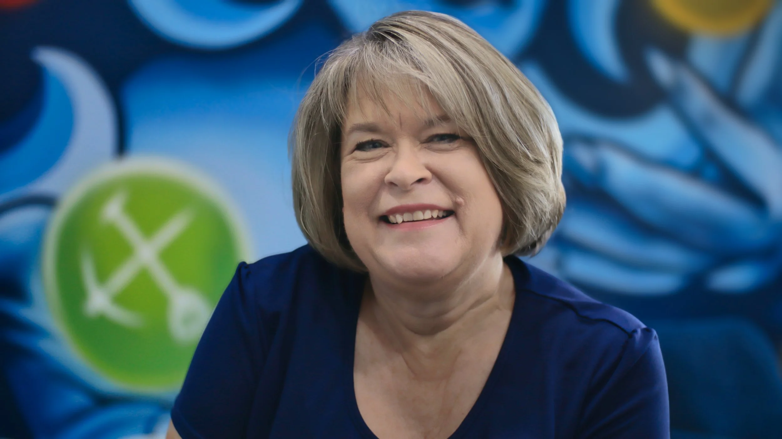 Senior Accountant, Cindy Reinders, smiling at camera in front of colourful blue and yellow mural. Located in Bunbury office.
