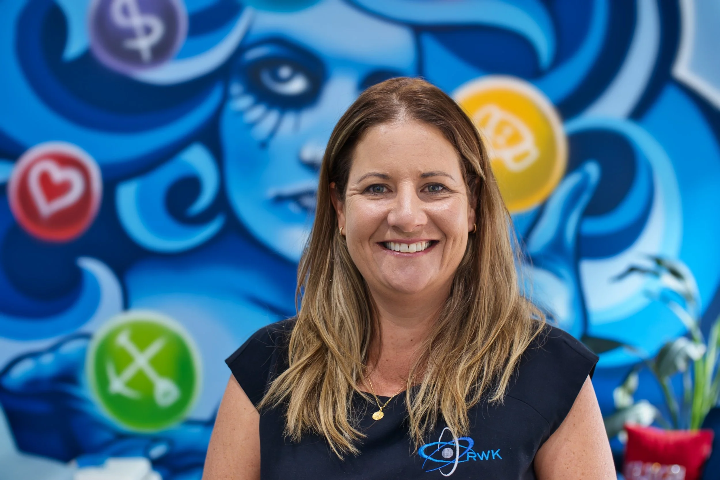 Director Gemma Denton, smiling at camera in front of colourful blue and yellow mural. Located in Bunbury office.