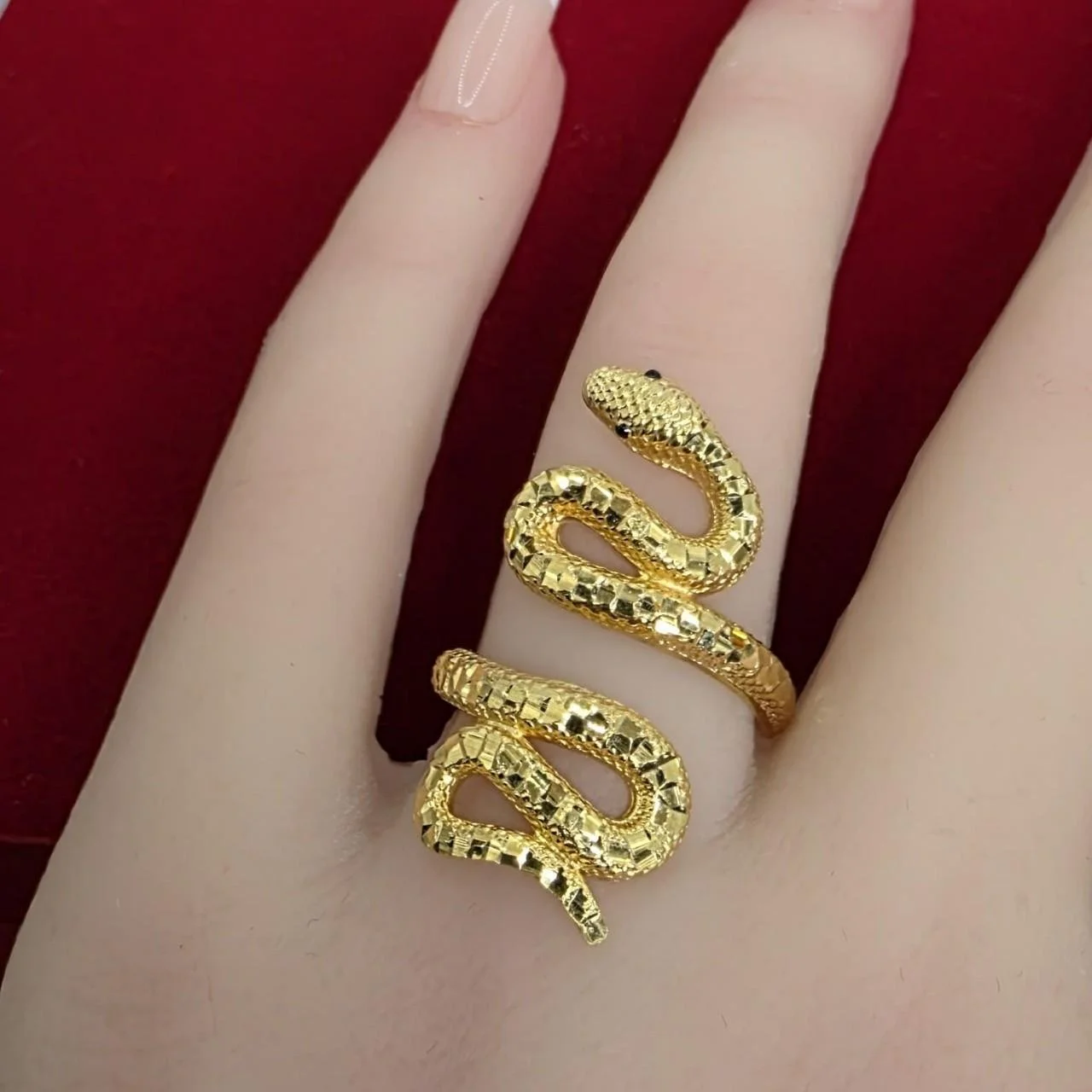 Gold snake-shaped ring with black eyes on a person's finger against a red background.
