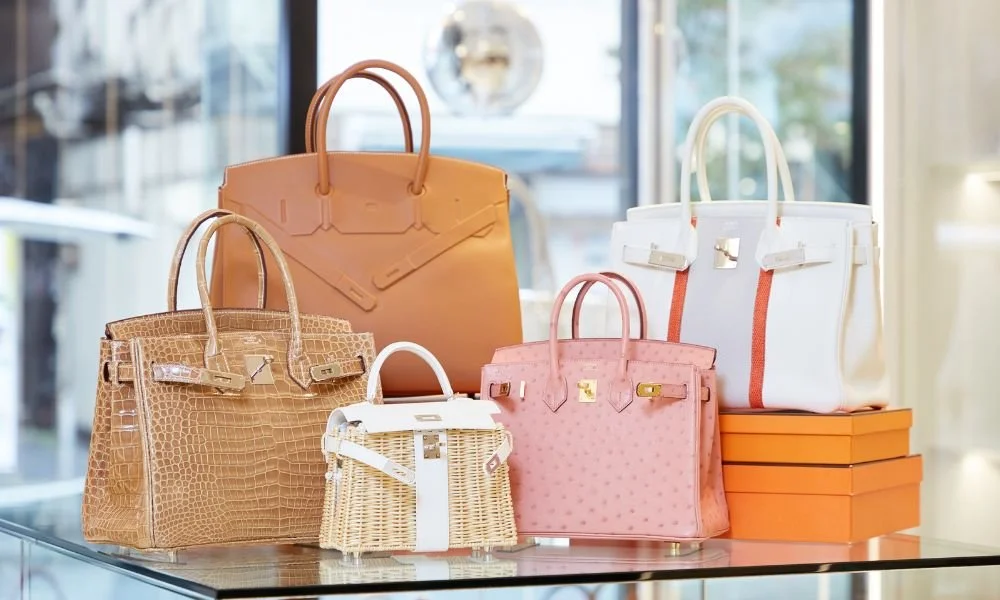 Display of five luxury handbags on a glass table in a store, featuring various colors and textures, with orange boxes beneath some of the bags.