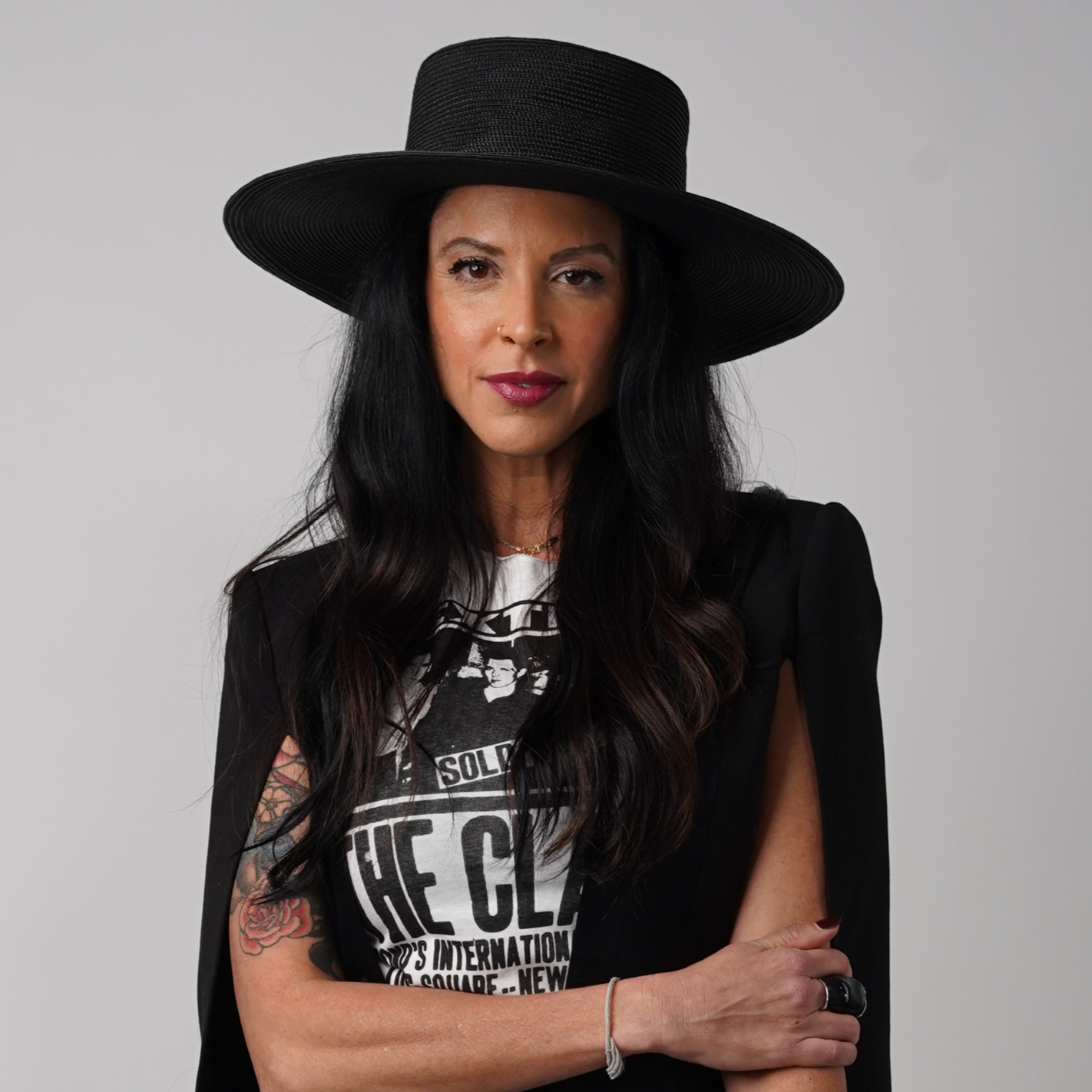 A woman with long dark hair wearing a black wide-brim hat and a dress with gold embroidery, smiling at the camera.